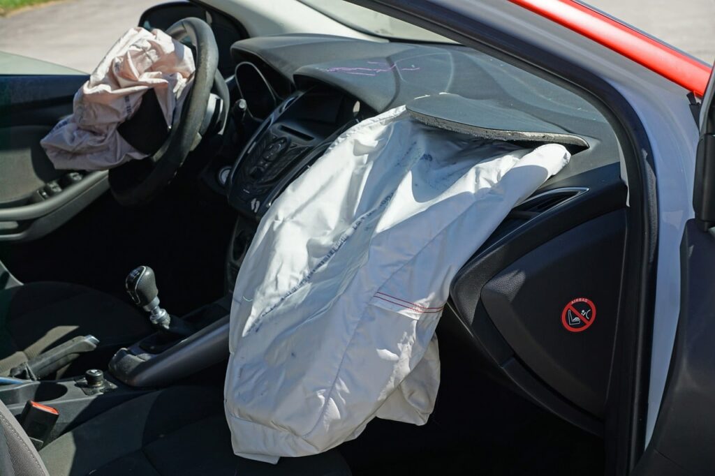 A car’s interior showing deployed airbags on the steering wheel and dashboard, indicating a recent collision. The front passenger seat is empty, and there is a no child seat sign on the dashboard.