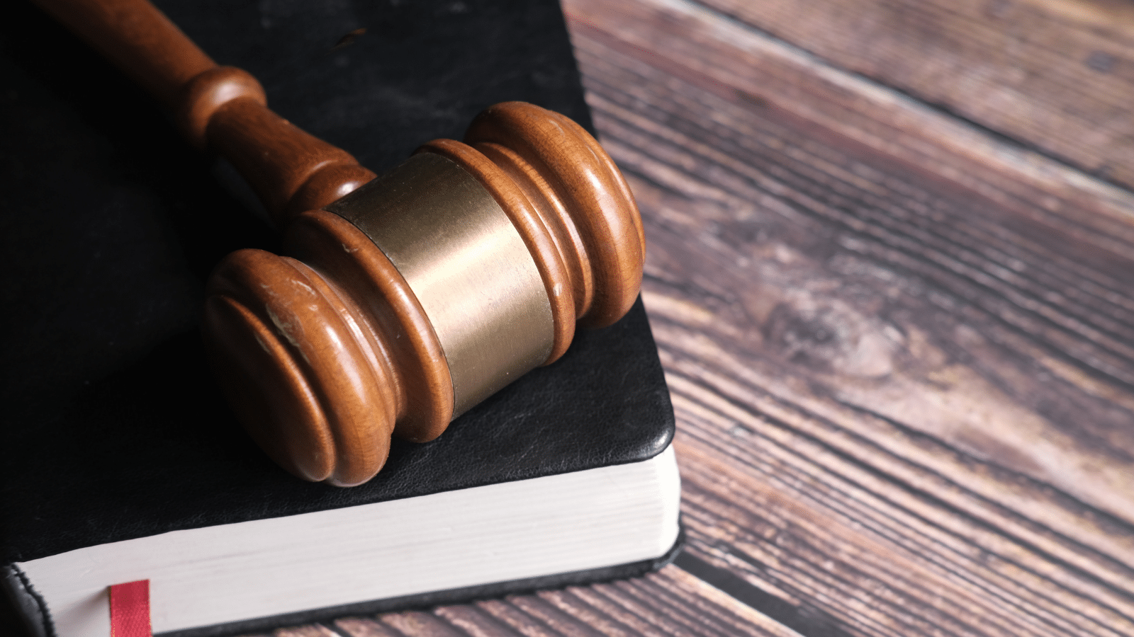 A wooden gavel rests on top of a closed black book with a white edge, placed on a rustic wooden table.