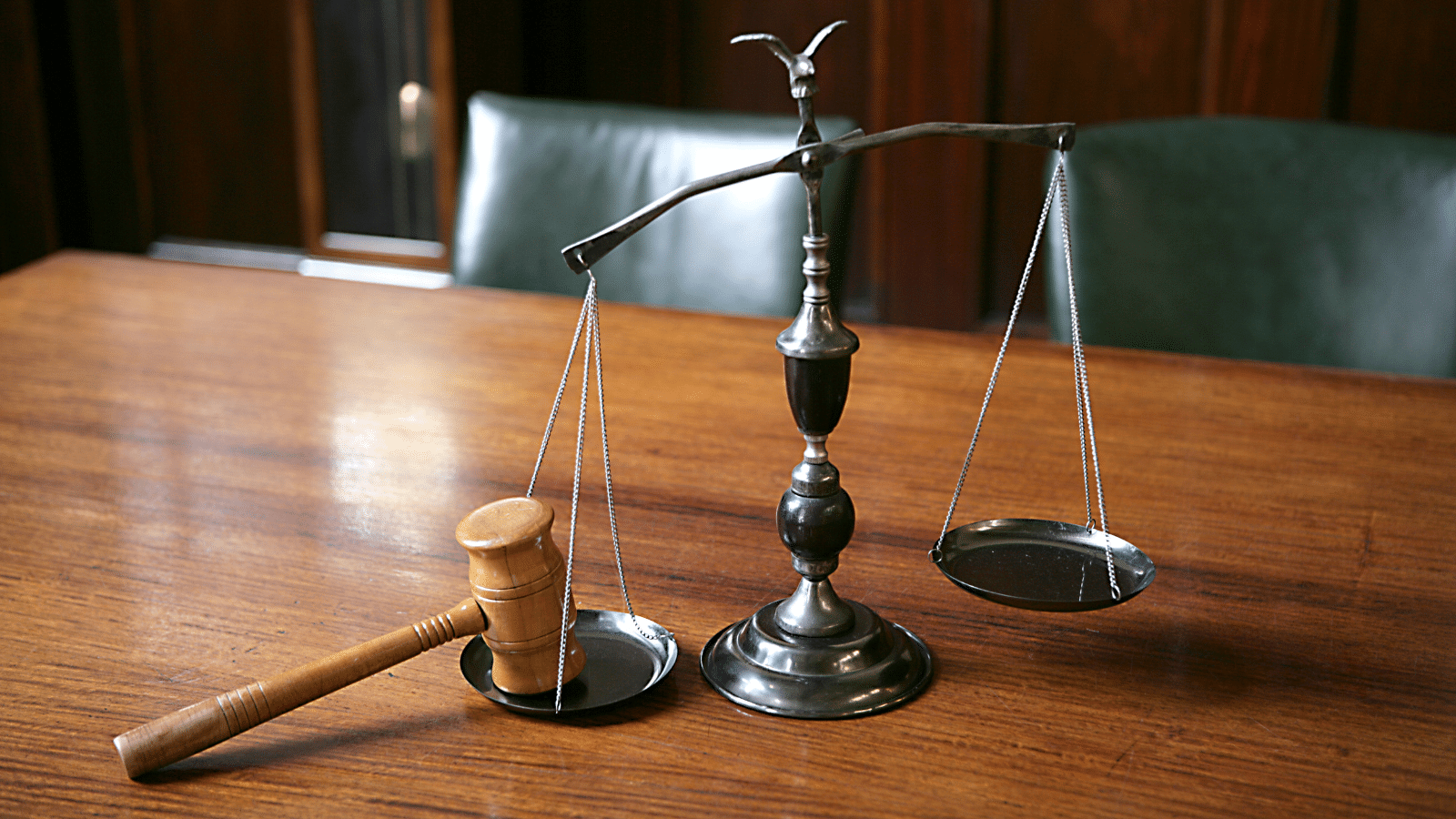 A brass balance scale on a wooden table, with a wooden gavel resting on one side of the scale, and the other side empty. Green leather chairs and wood paneling are in the background.