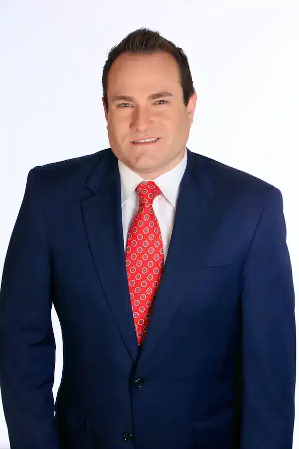 A man wearing a dark blue suit, white dress shirt, and a red patterned tie stands against a plain white background, smiling at the camera.