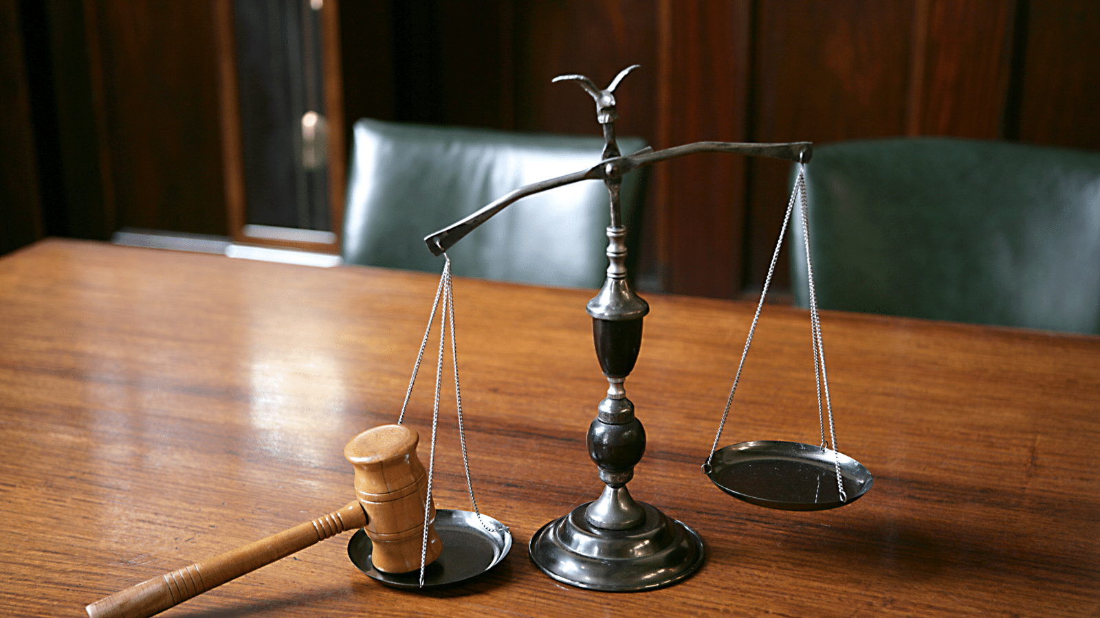 A wooden gavel and a set of metal balance scales sit on a polished wooden table in a courtroom setting, with a green leather chair and wood-paneled wall in the background.
