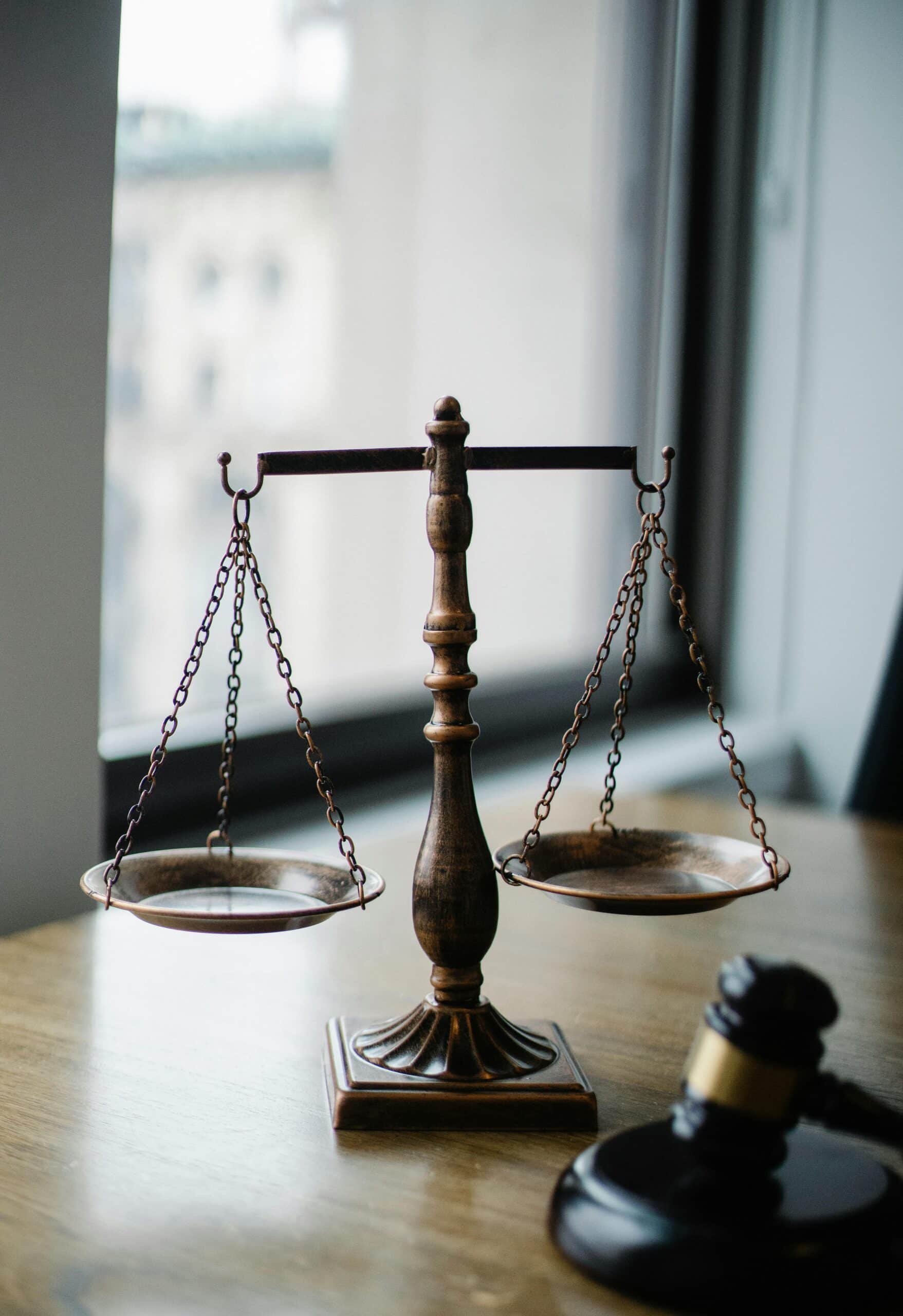 A bronze balance scale and a wooden judge’s gavel rest on a wooden desk near a window, symbolizing justice and law.
