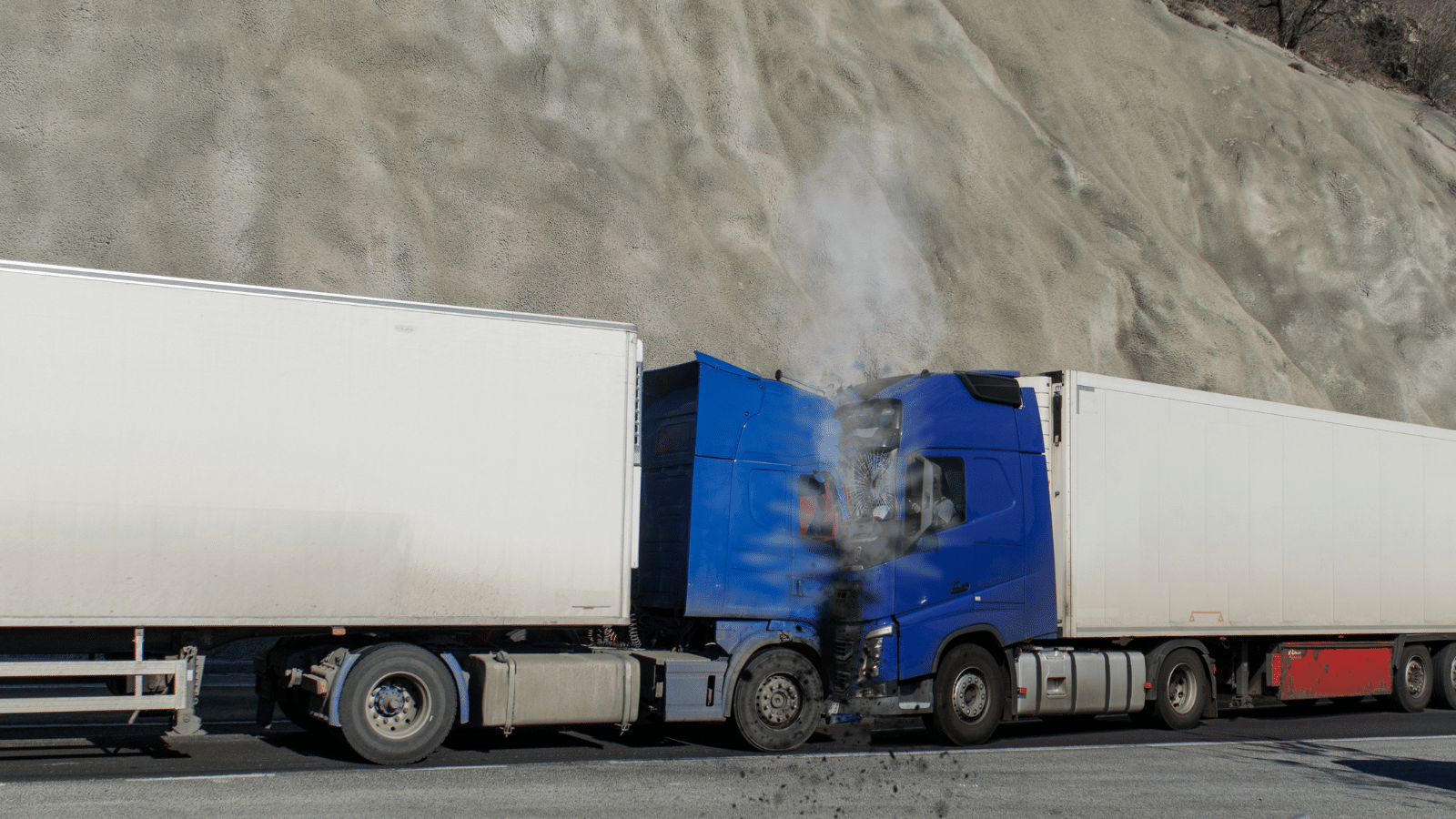 A blue semi-truck with a damaged front has crashed into the rear of a white trailer on a road beside a steep, rocky hillside. Smoke is rising from the blue trucks engine area.