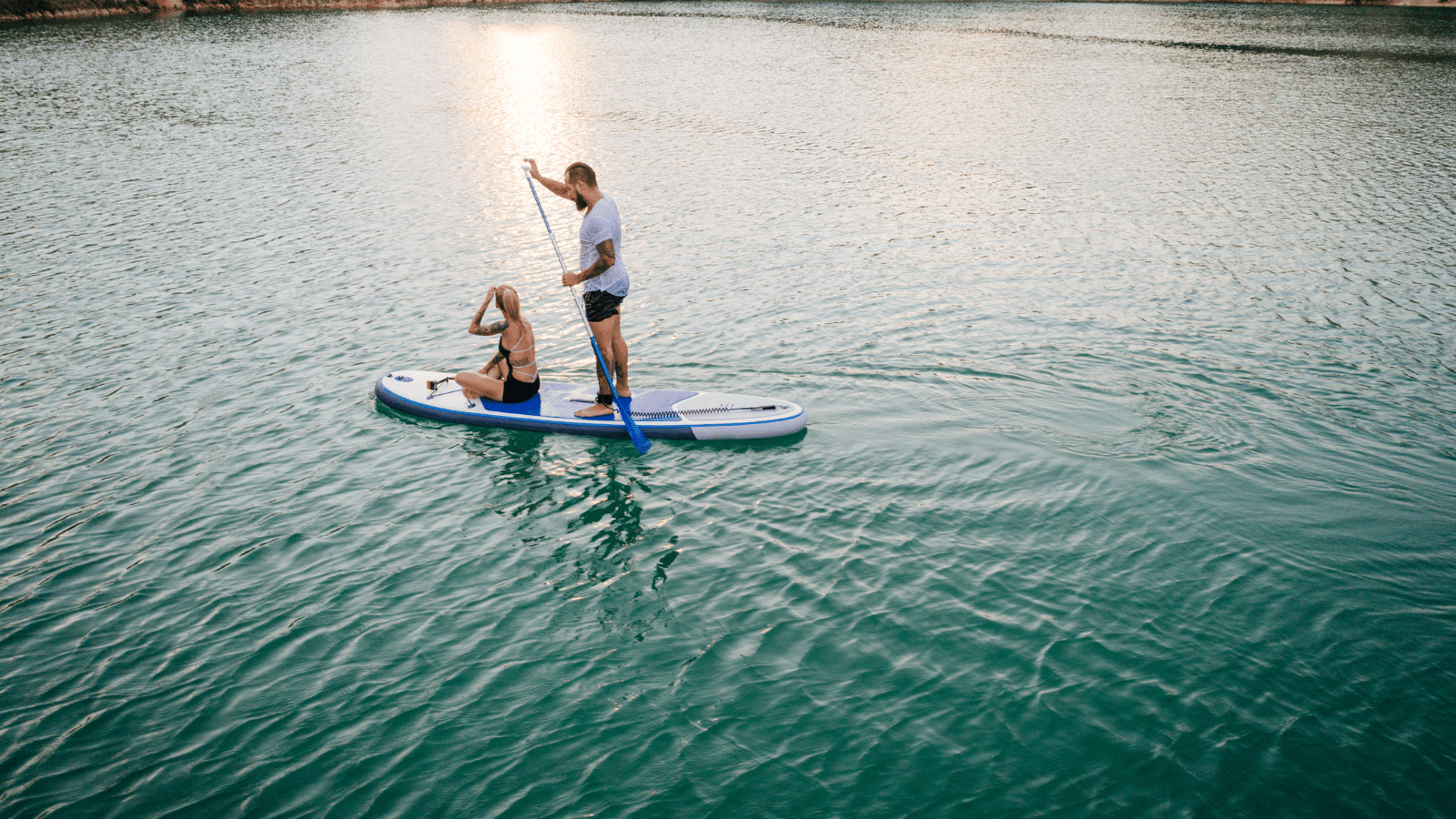 Two people paddleboarding on calm turquoise water at sunset; one person is sitting while the other stands and paddles.