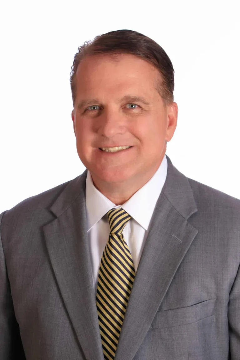 A middle-aged man with short brown hair, wearing a gray suit, white shirt, and yellow-and-black striped tie, smiles at the camera against a plain white background.