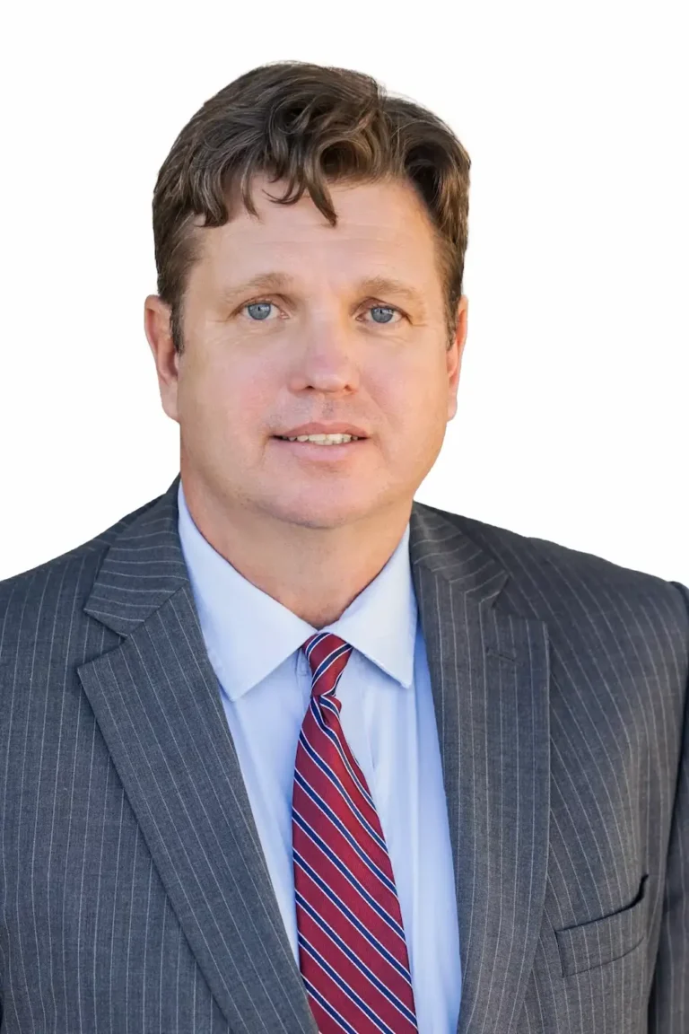 A man with short brown hair wearing a gray pinstripe suit, light blue dress shirt, and red striped tie, poses for a professional headshot against a white background.