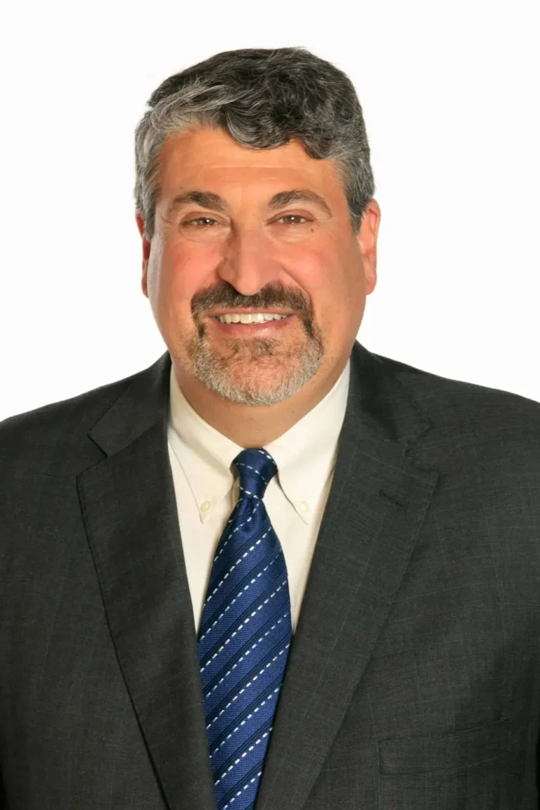 A middle-aged man with short, curly gray hair and a beard, wearing a dark suit, white shirt, and blue striped tie, smiling at the camera against a white background.