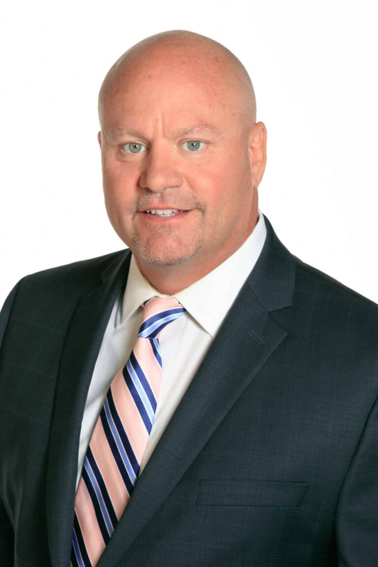 A bald man in a dark suit, white shirt, and striped tie poses against a plain white background, looking directly at the camera with a neutral expression.