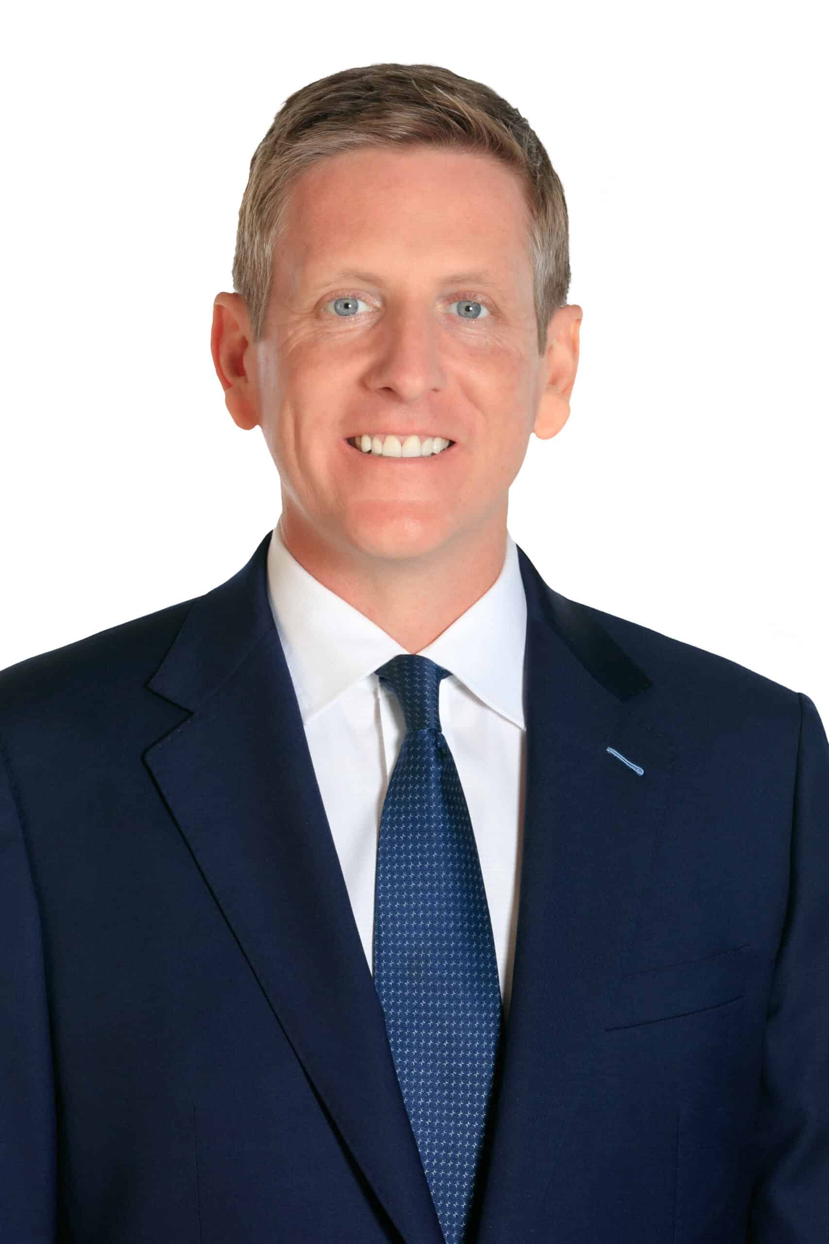 A man in a dark blue suit, white shirt, and blue tie smiles at the camera against a white background.