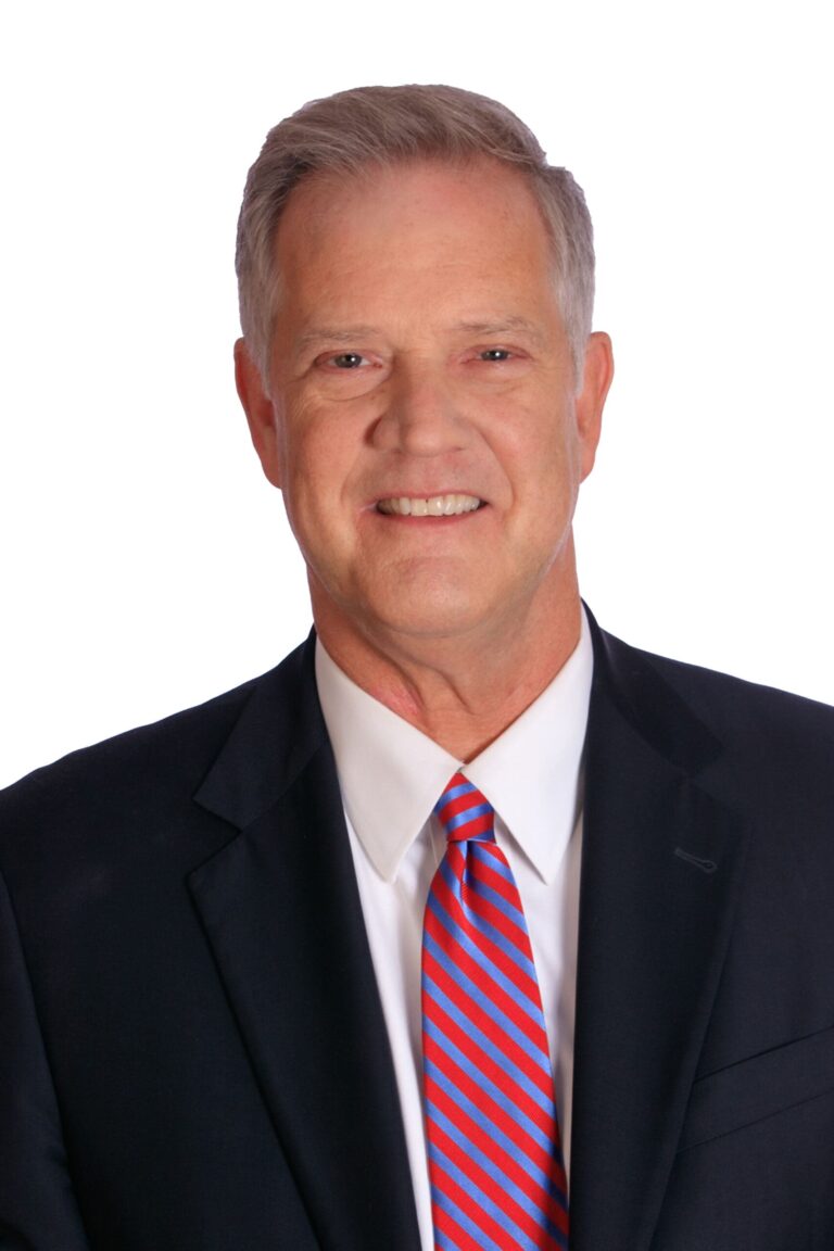 Thomas M. Burke, a middle-aged man with short gray hair, wearing a dark suit, white shirt, and red-and-blue striped tie, smiles at the camera against a plain white background.