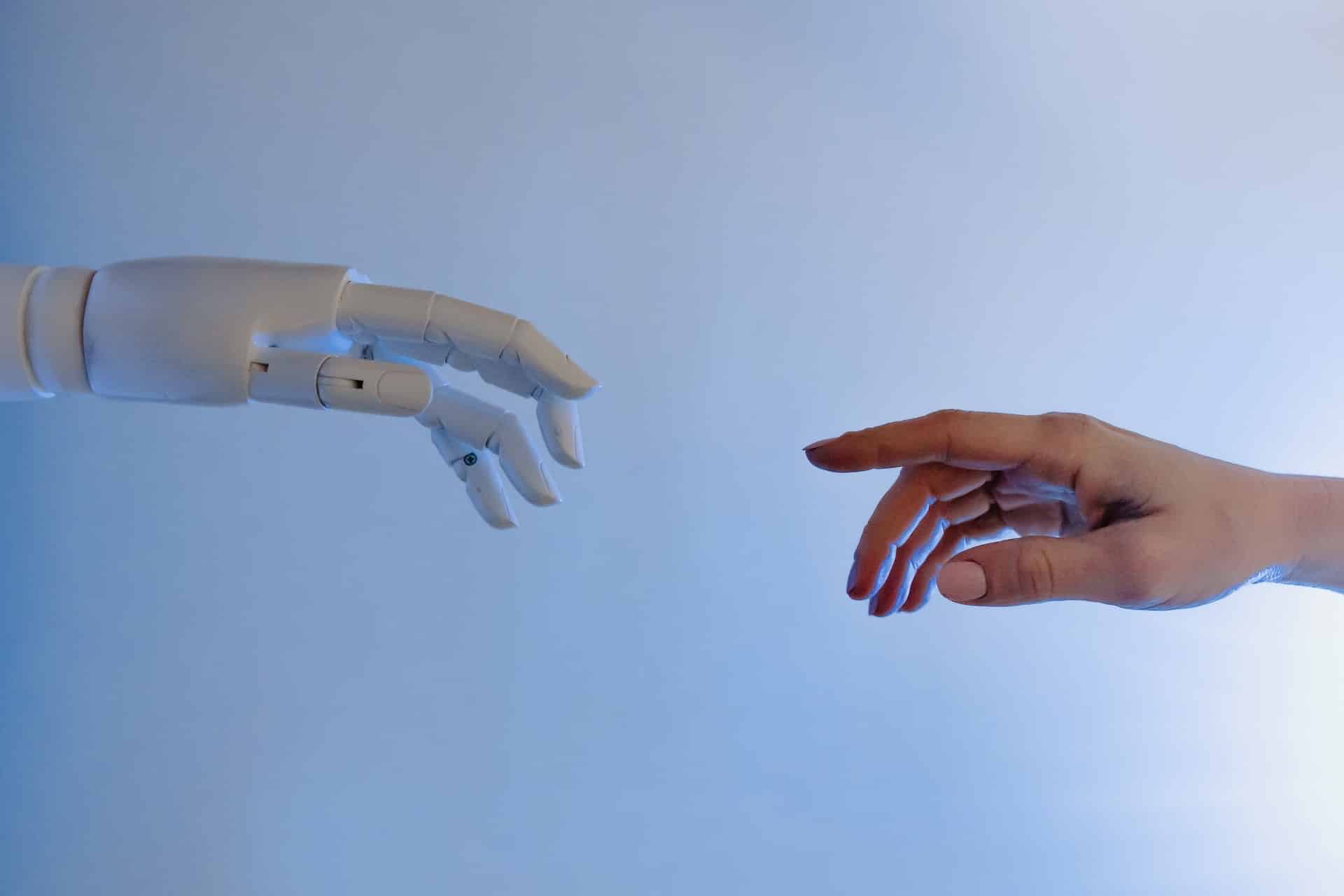 A robotic hand and a human hand reach toward each other against a light blue background, with fingertips almost touching, symbolizing the connection between technology and humanity.