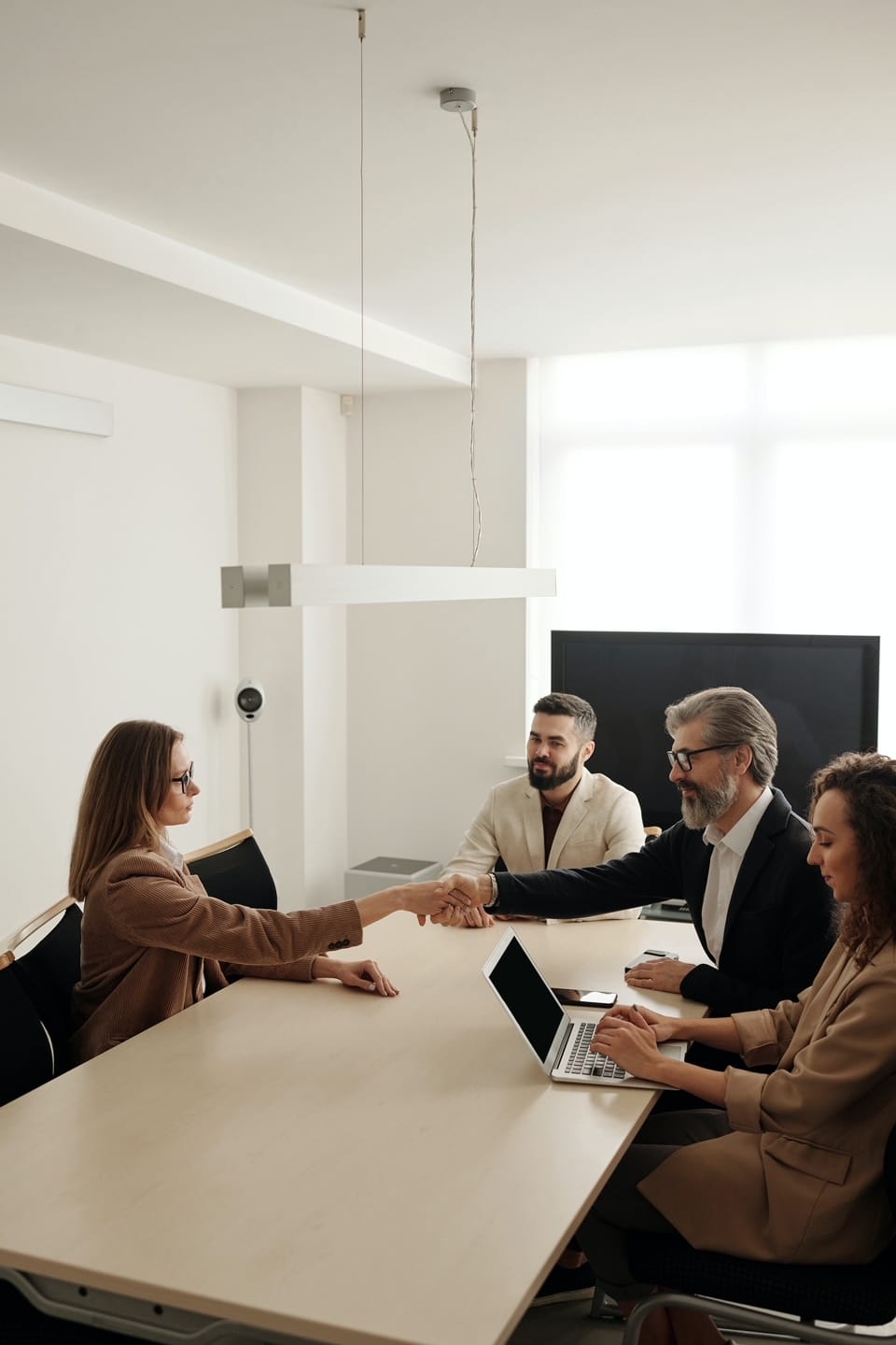 A woman is shaking hands with a man across a conference table during a meeting or interview, while two other people sit beside him, one using a laptop. The setting is a modern, well-lit office.