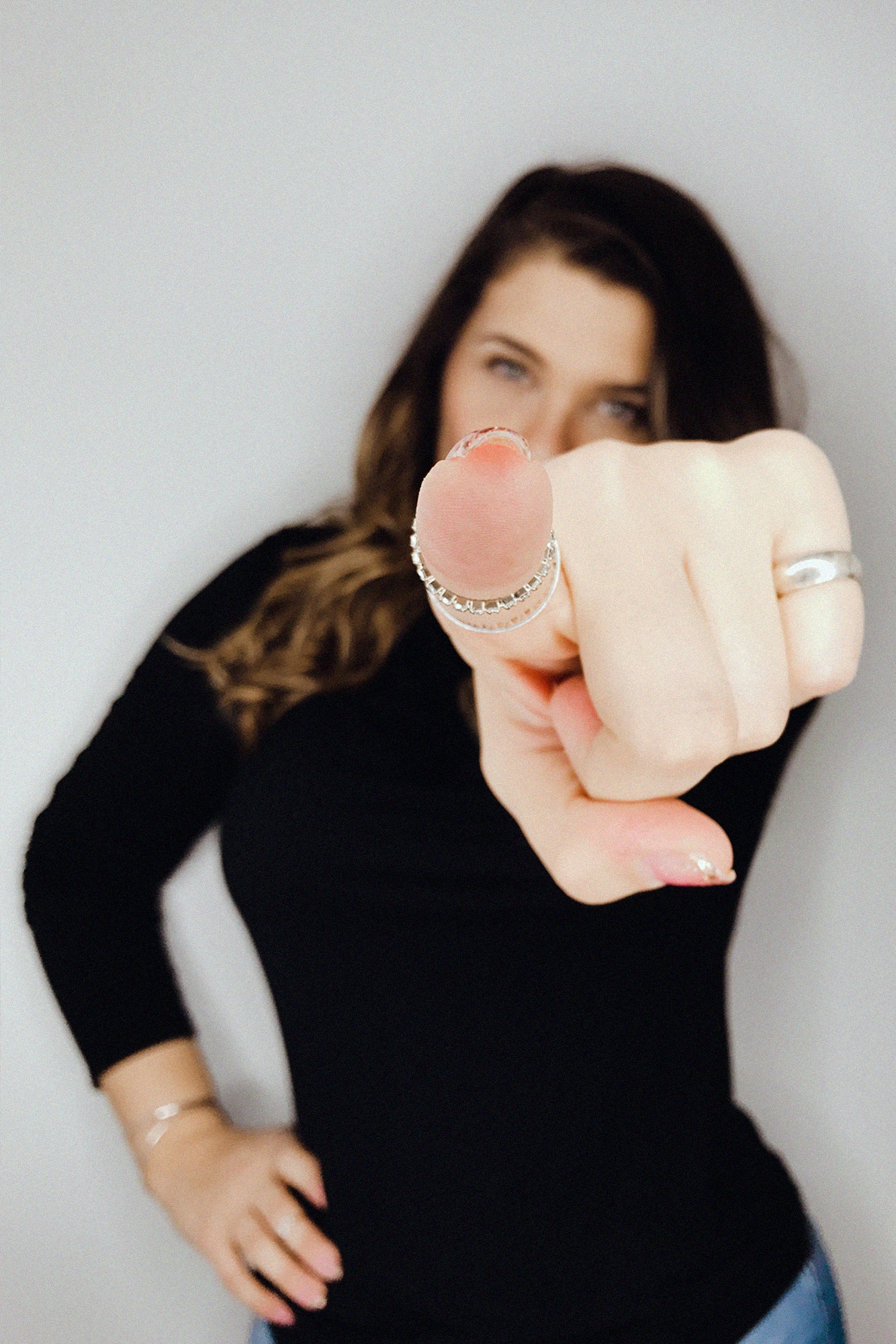 A woman with long brown hair wearing a black top points directly at the camera. Her face is out of focus while her hand, adorned with rings, is in sharp focus. She stands against a plain light background.