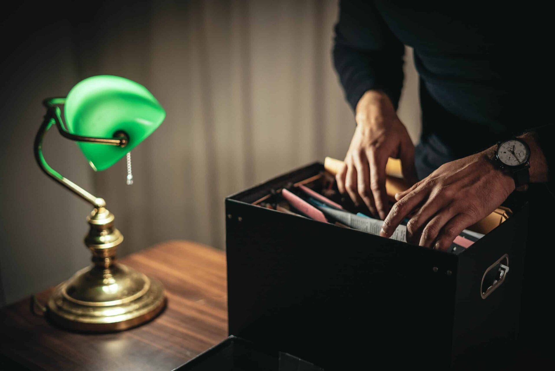 A person wearing a watch searches through files in a black box on a wooden desk next to a green-shaded brass lamp.