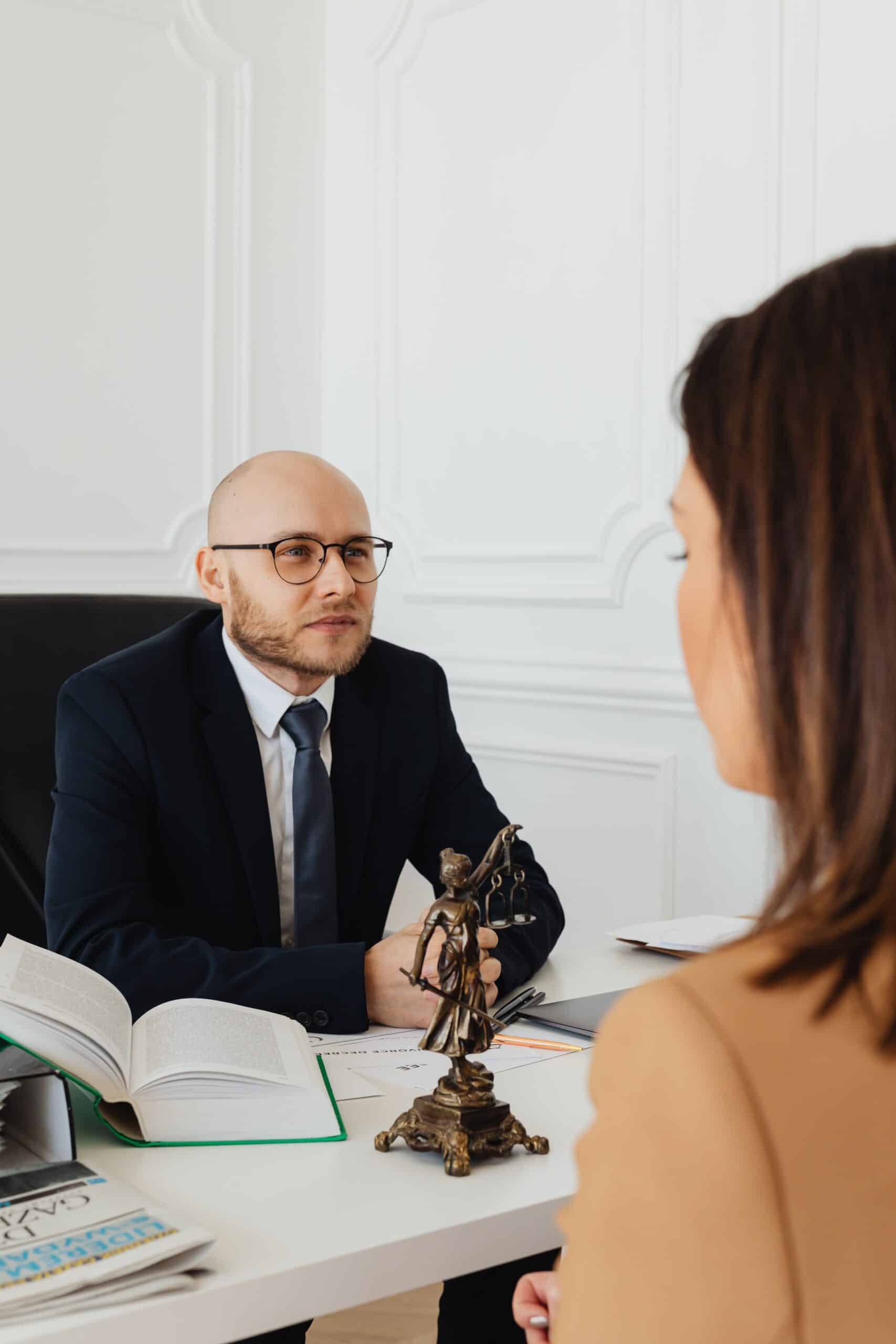 A man in a suit and glasses sits at a desk with open books and a Lady Justice statue, speaking to a woman seated across from him in an office setting.