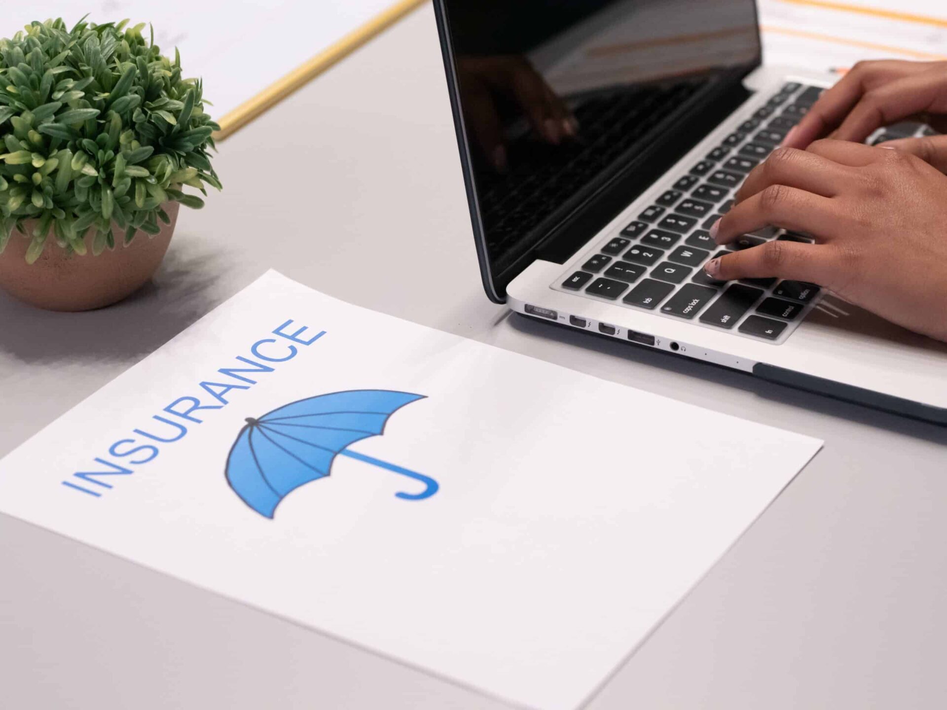 A person typing on a laptop next to a document labeled INSURANCE with a blue umbrella illustration, and a small potted plant on the desk.