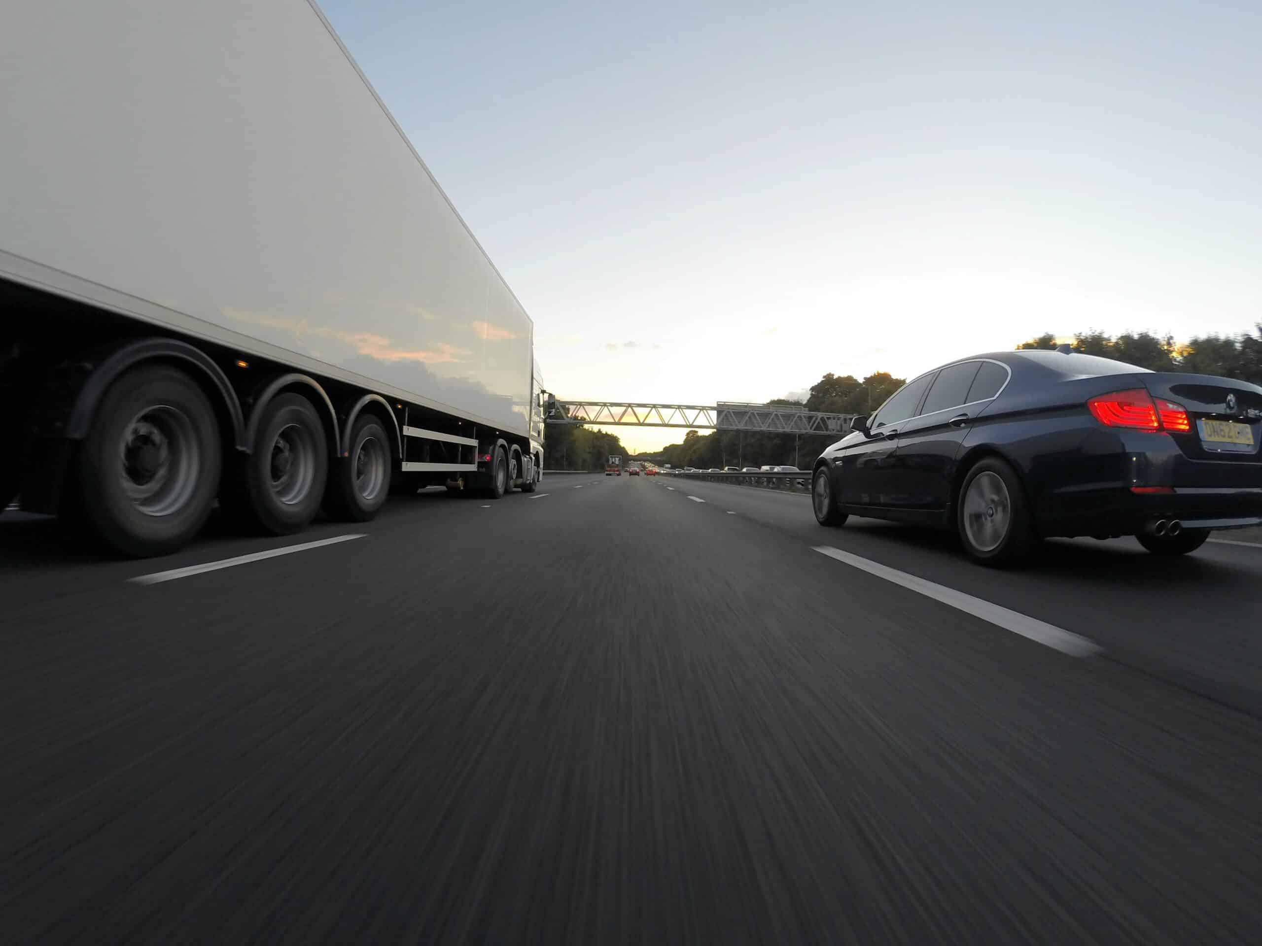 A black car and a large white truck drive side by side on a multi-lane highway at sunset, with a pedestrian bridge ahead and trees lining the road. The  is taken from a low perspective close to the ground.