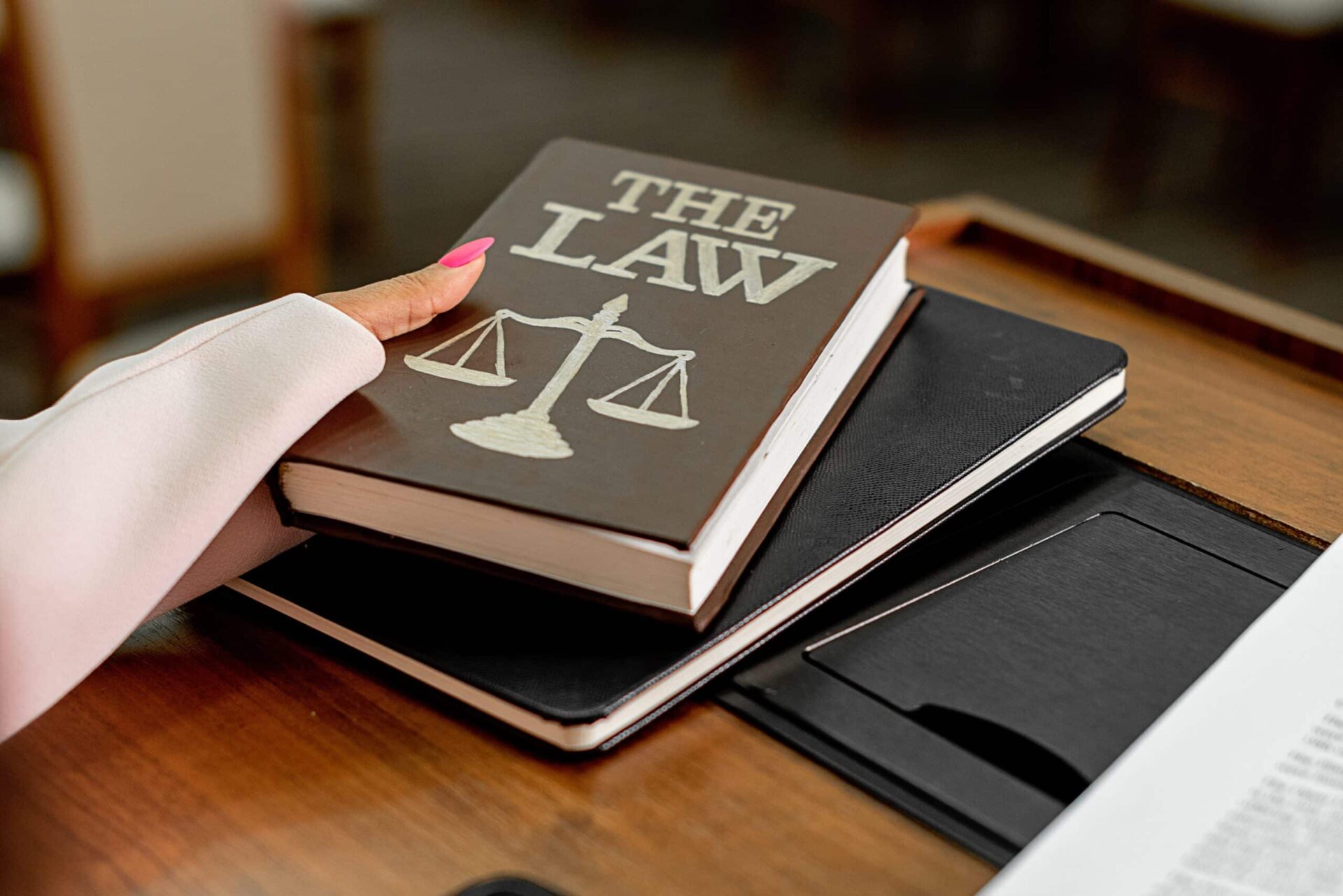 A person with pink nail polish holds a book titled THE LAW with a scale symbol on the cover, resting on top of a closed notebook on a wooden desk.