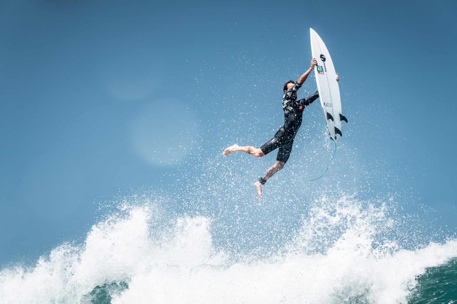 A surfer in a black wetsuit is airborne above a wave, reaching up and holding their surfboard with one hand as water sprays up around them against a clear blue sky.