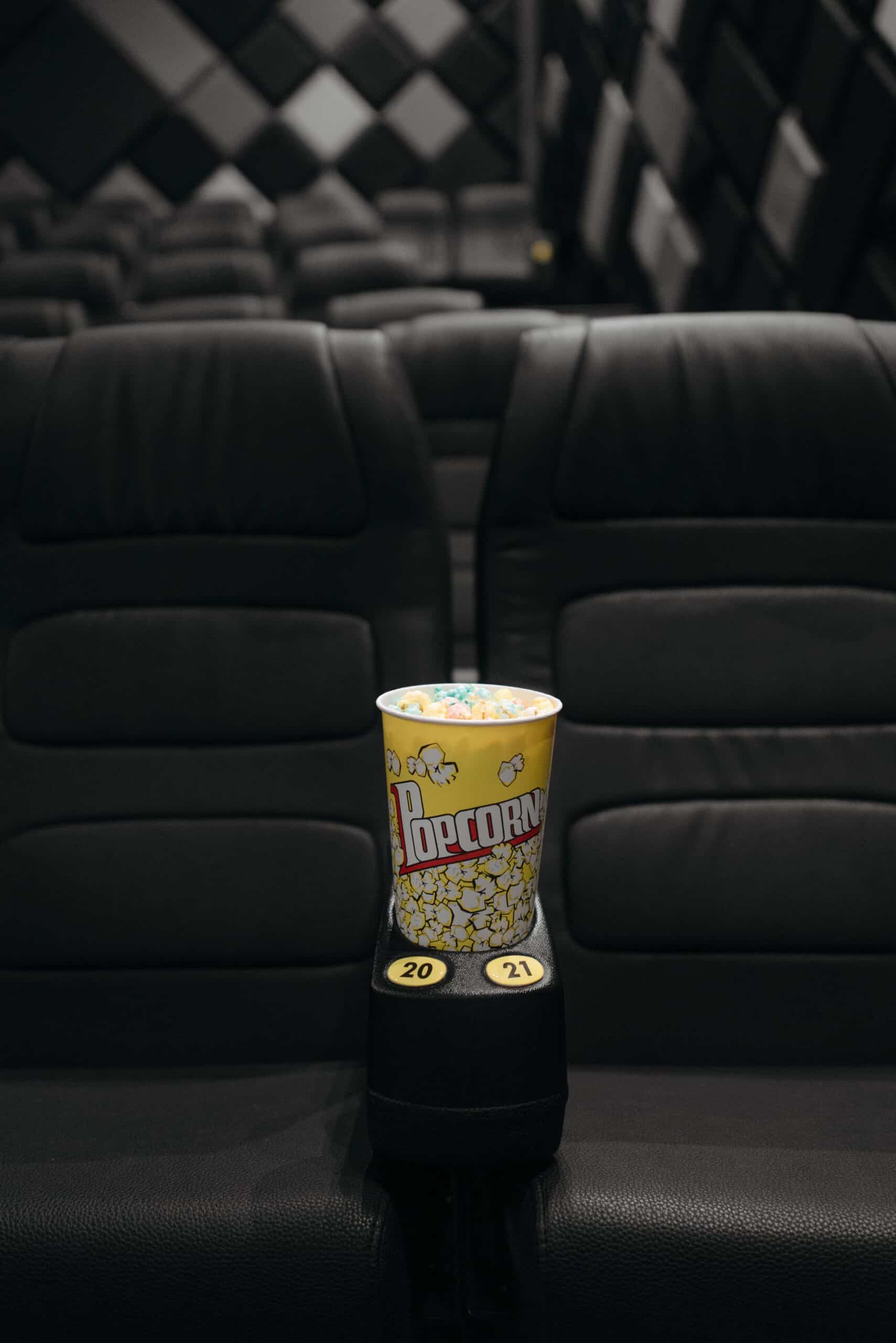 A cup of popcorn sits on an armrest between two empty black cinema seats, marked with seat numbers 20 and 21, in a dimly lit movie theater.