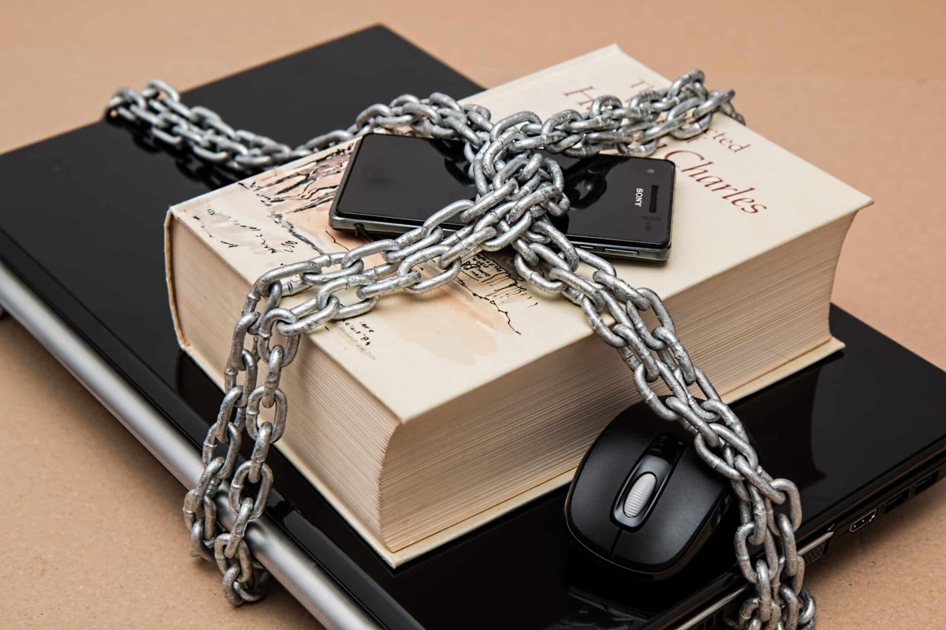 A thick book, closed laptop, smartphone, and computer mouse are stacked together, secured tightly with a heavy metal chain.