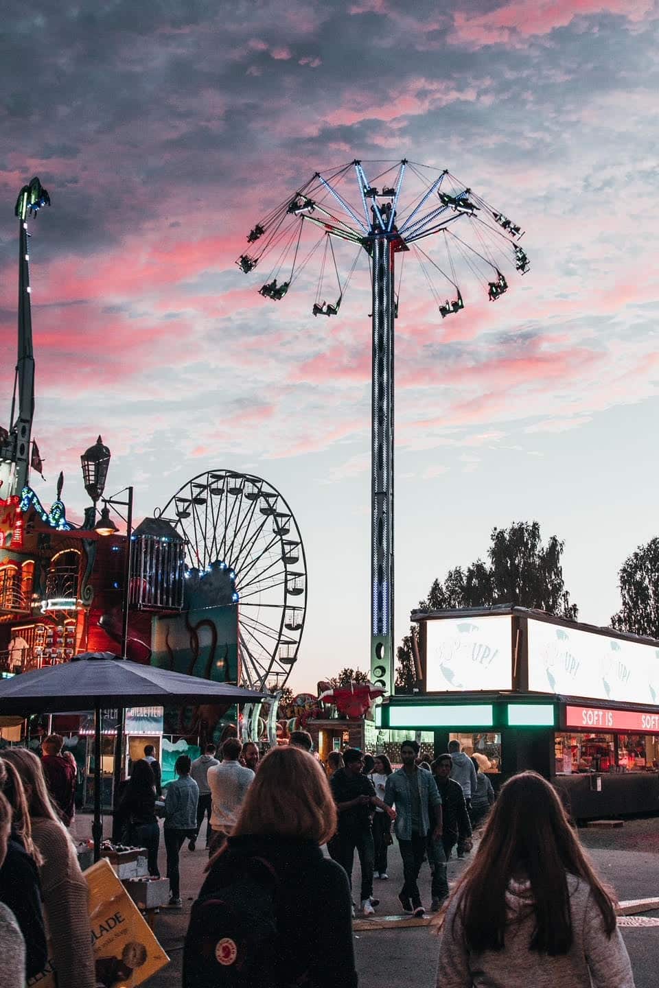 People walk through a lively fairground at sunset, with a tall swing ride in motion, a Ferris wheel, colorful lights, and a few food stands visible under a pink and purple sky.