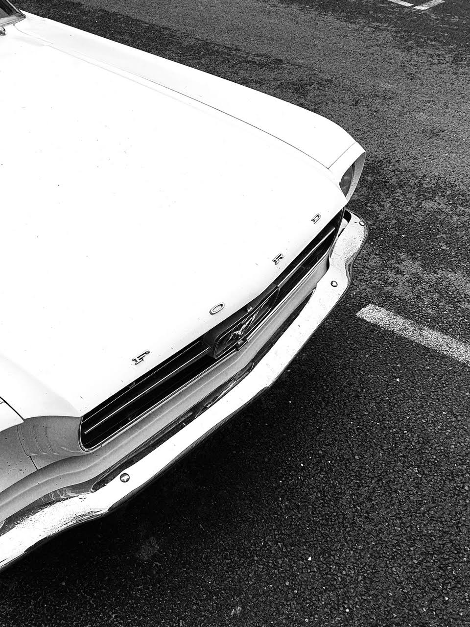 Black and white  showing the front end of a classic white Ford Mustang parked on an asphalt surface, with part of the hood, grille, and emblem visible.