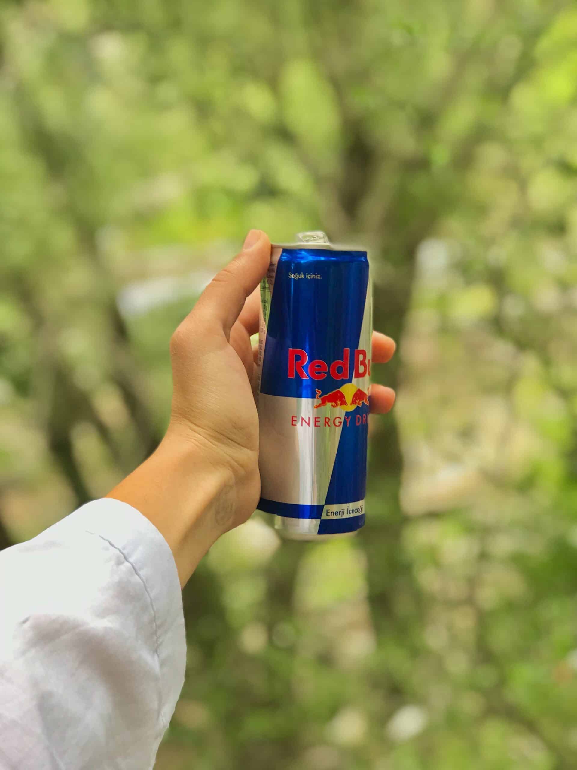 A person wearing a white sleeve holds a can of Red Bull energy drink outdoors, with a blurred background of green trees.