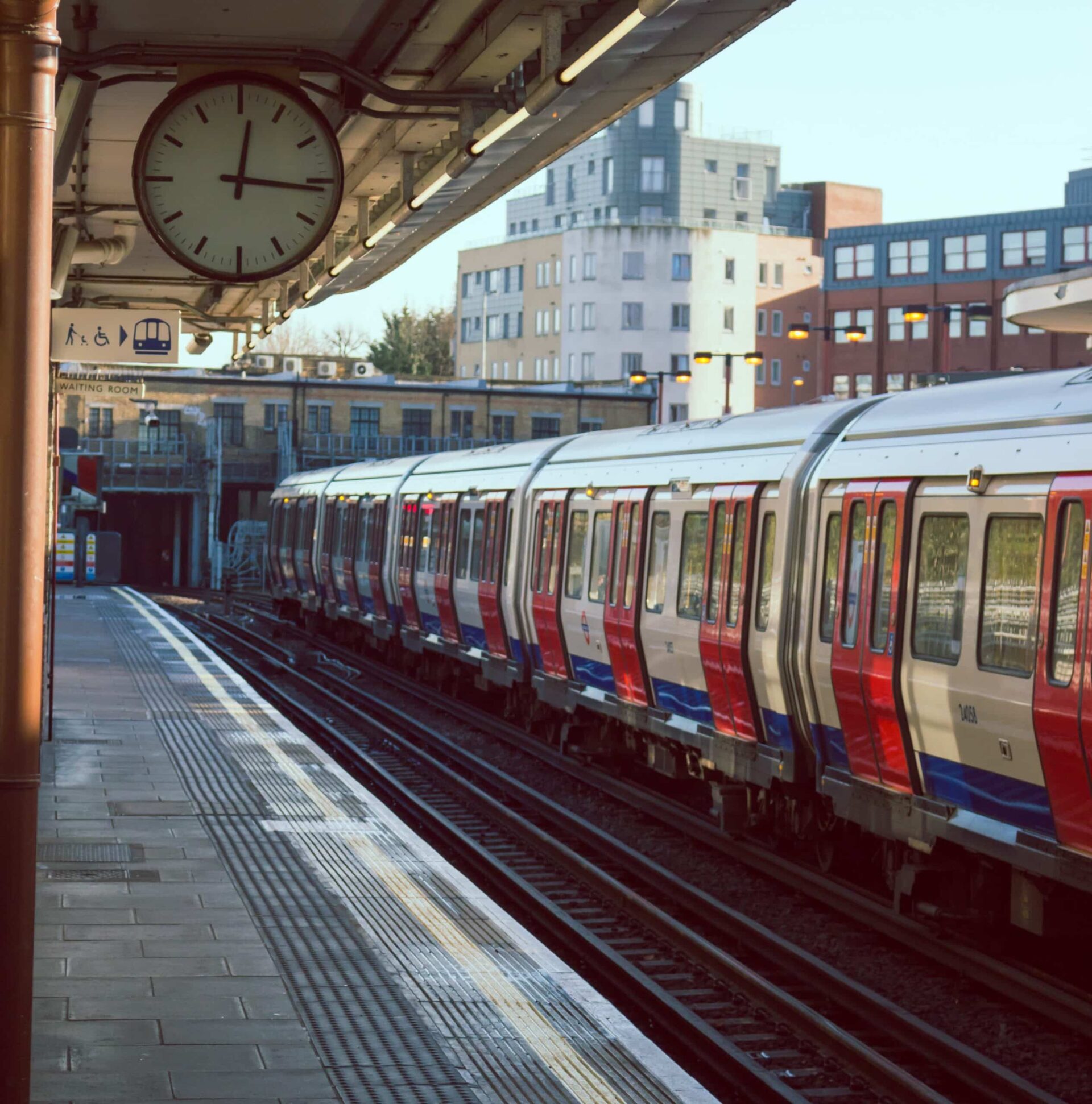 A London Underground train with red, white, and blue carriages is stopped at an outdoor station platform, with a clock showing 8:53, buildings, and a sign with a train symbol in the background.