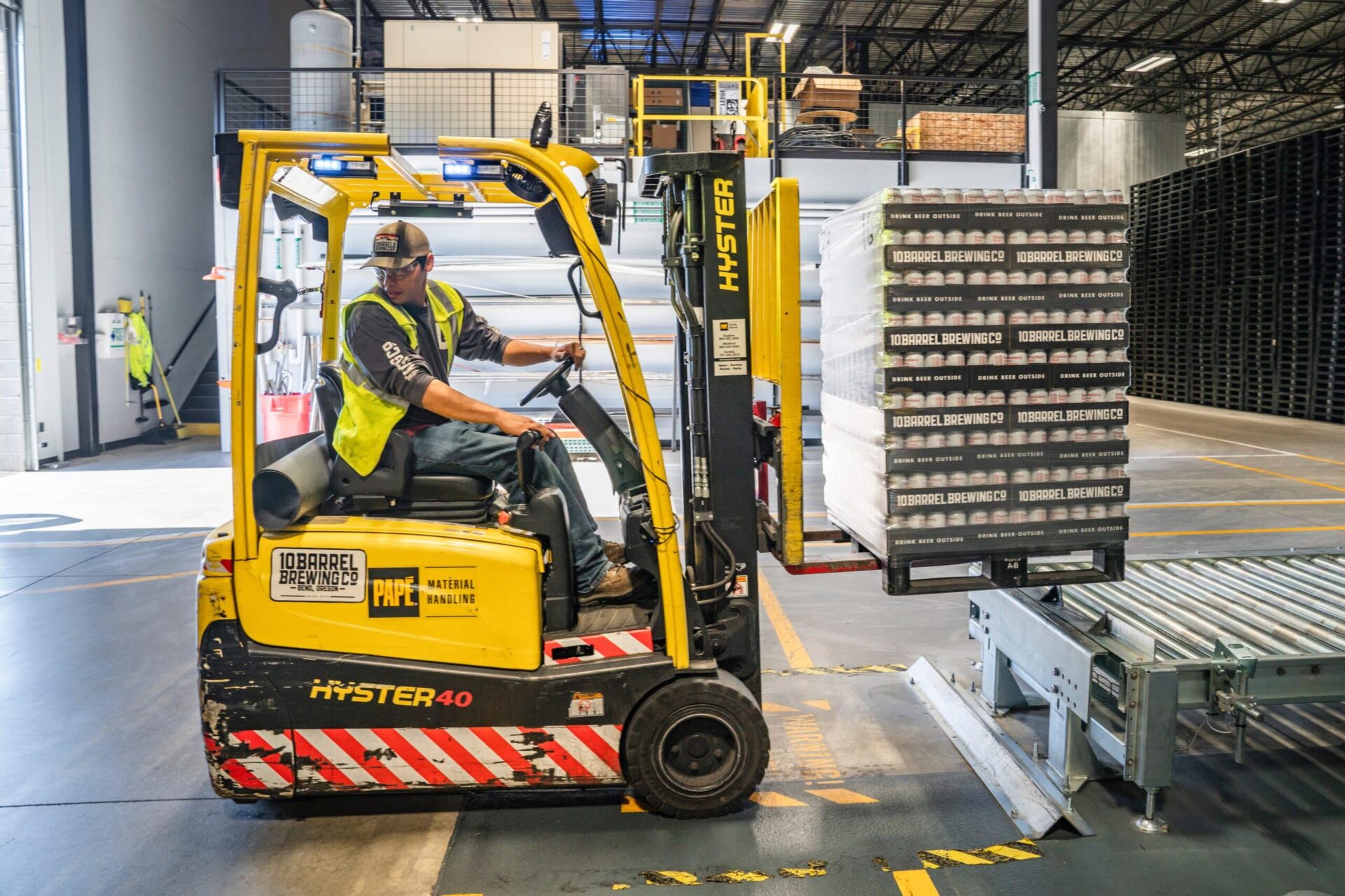 A worker in a safety vest and cap operates a yellow forklift, moving a pallet of wrapped goods in a warehouse with high ceilings and industrial equipment.