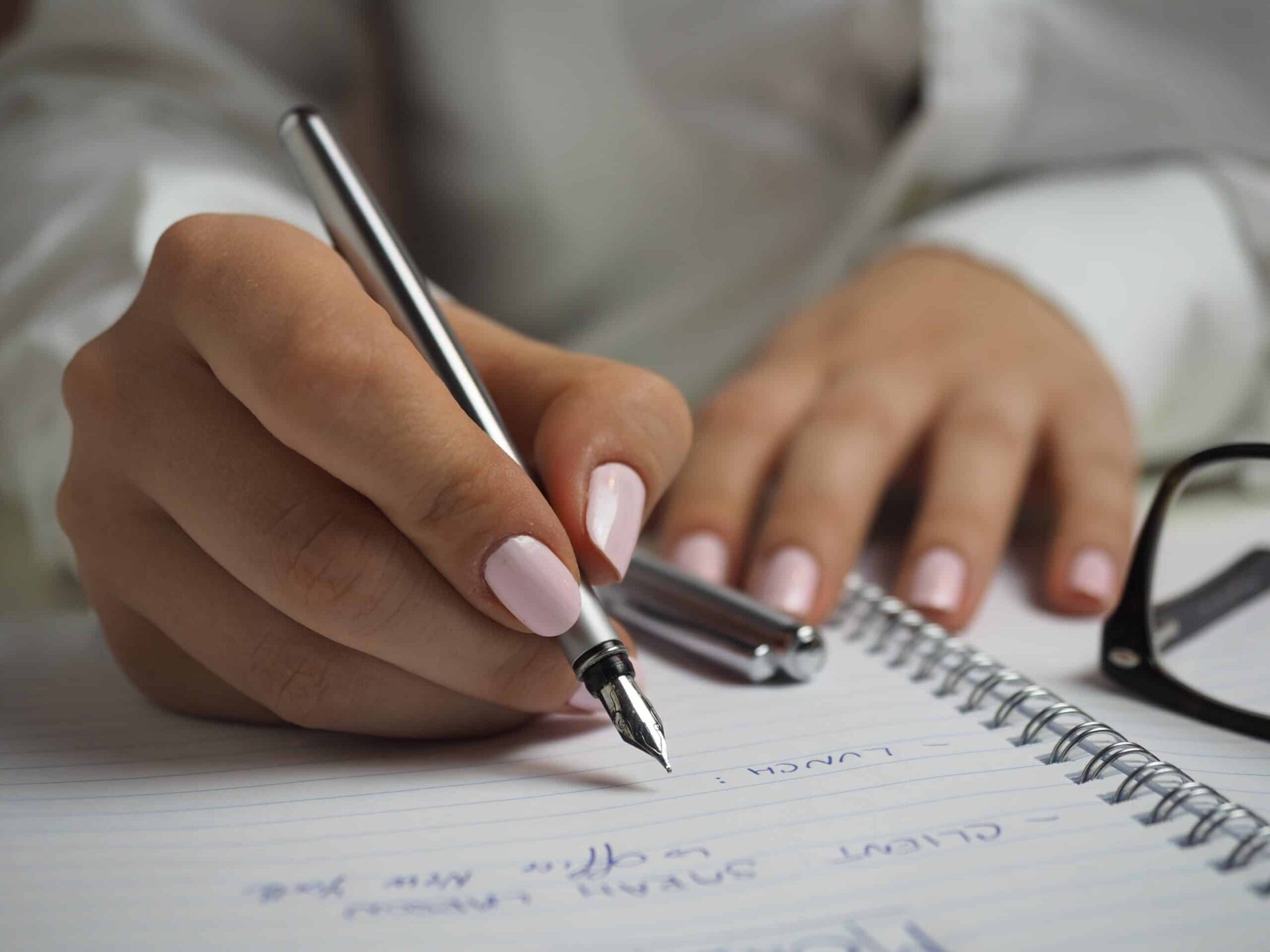 A person with light pink nail polish writes with a silver fountain pen on a spiral notebook. Their other hand rests on the page, and a pair of black eyeglasses lies nearby.