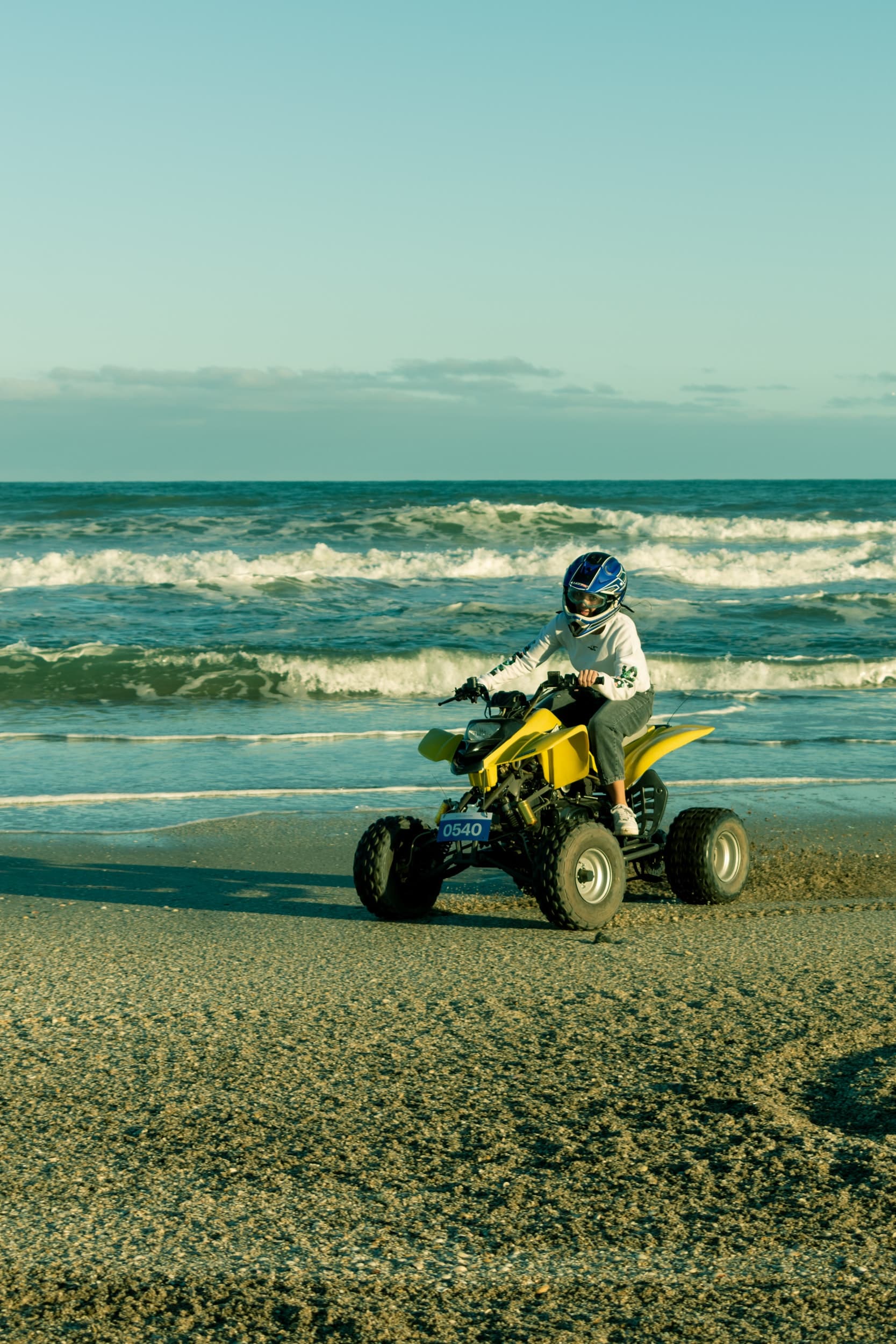 A person wearing a helmet and hoodie rides a yellow ATV along the shore of a sandy beach, with waves and the ocean visible in the background under a clear sky.