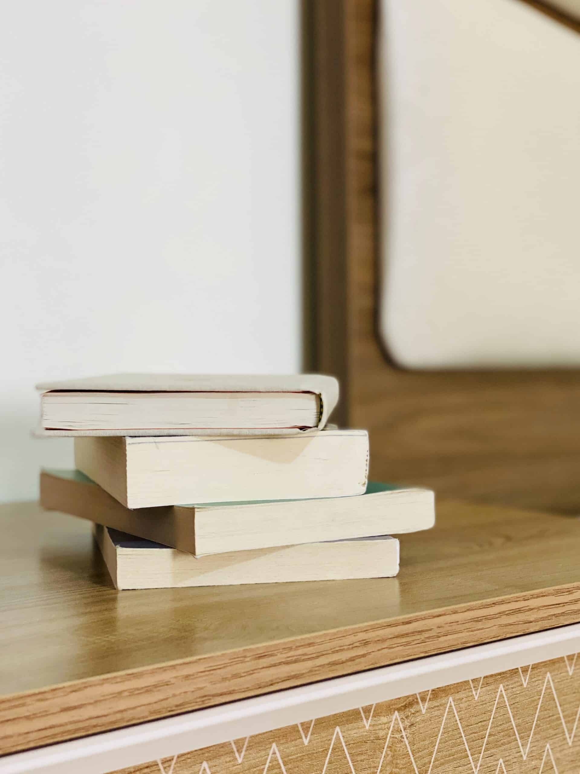 A small stack of three closed books sits on a wooden surface in front of a mirror with a brown frame. The background features light-colored walls and part of a patterned dresser.