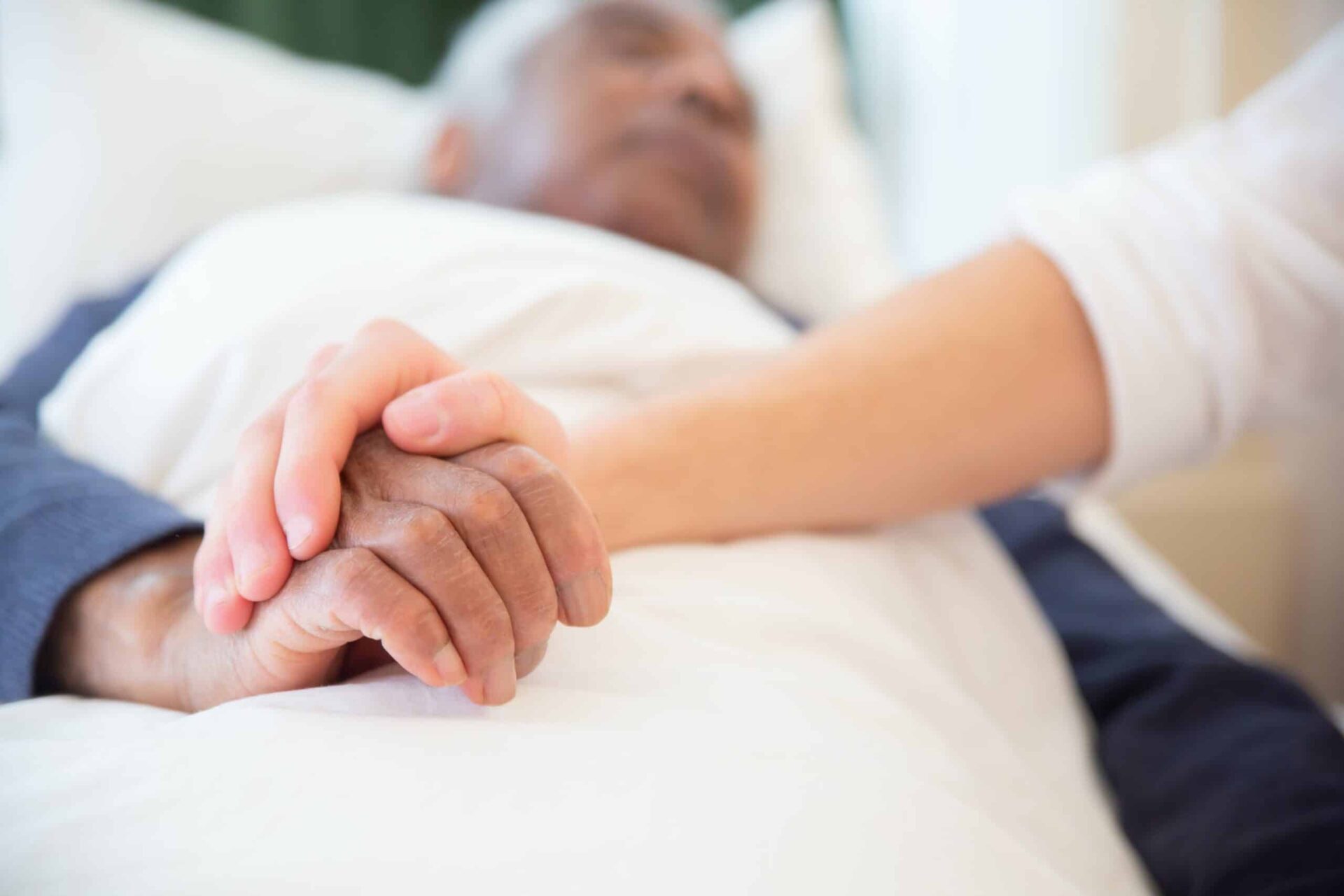 A close-up of two people holding hands, with one person lying in a hospital bed and the other offering comfort and support.