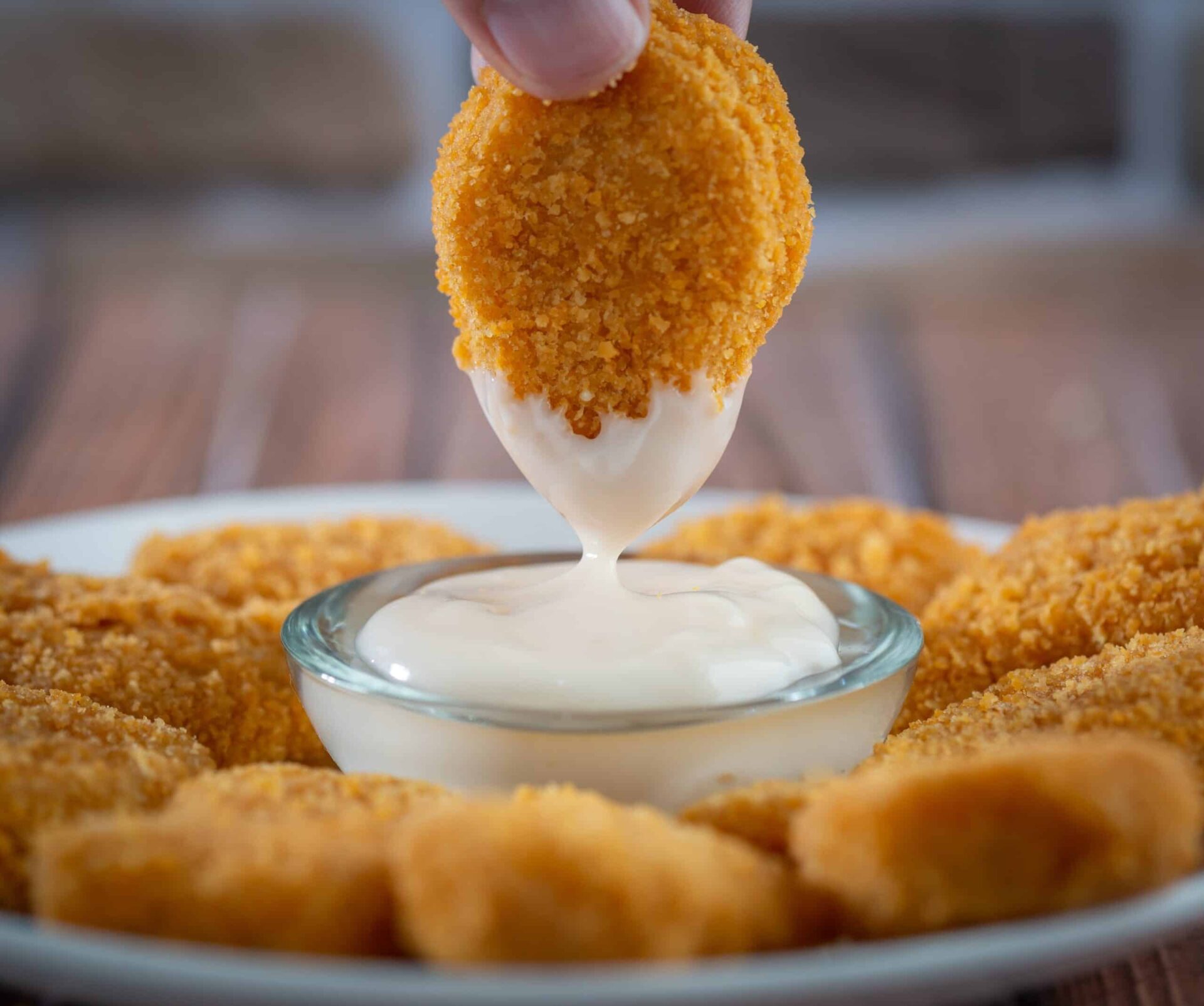 A hand dips a crispy, breaded chicken nugget into a small bowl of creamy white sauce, with more nuggets arranged on a plate in the background.