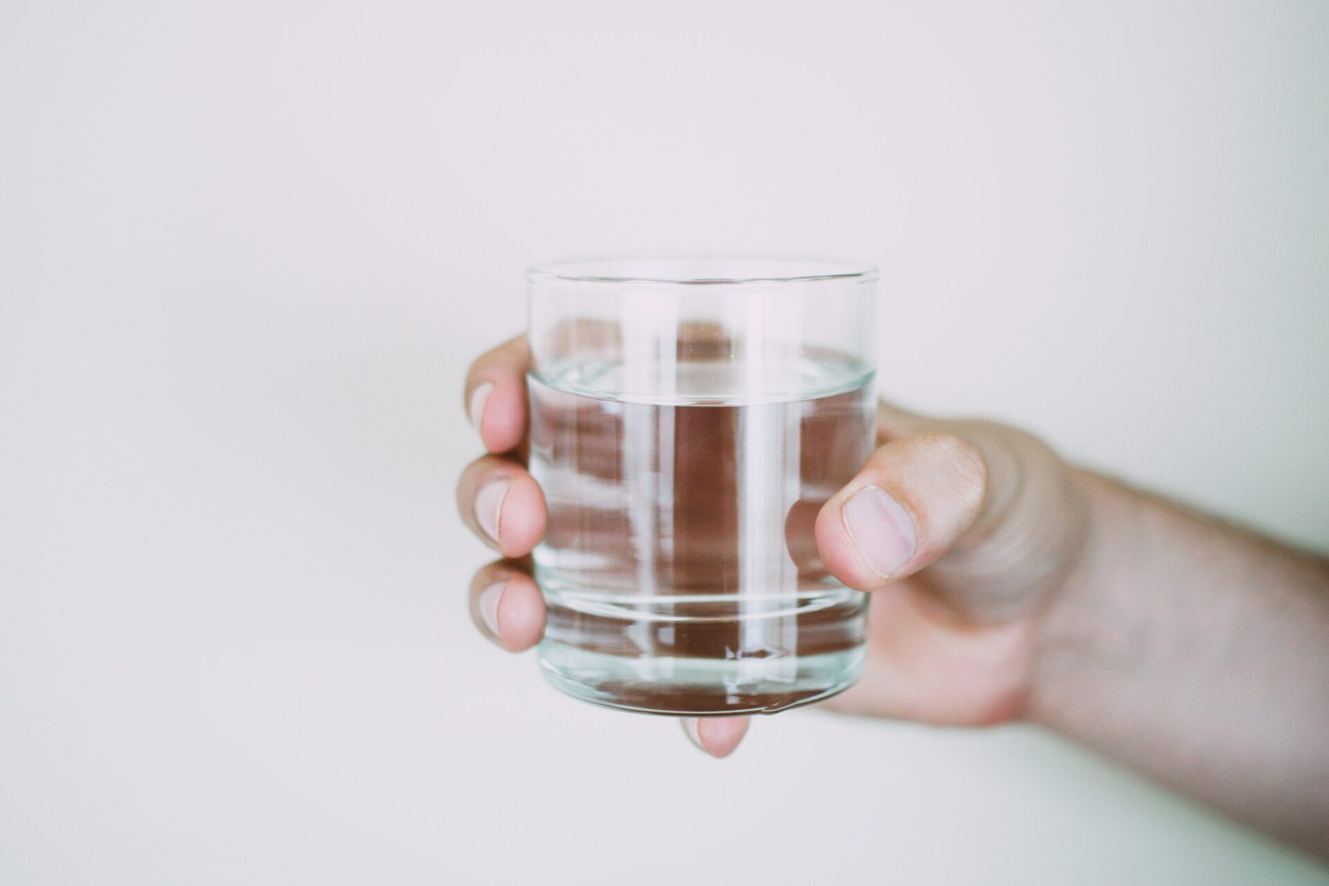 A hand holding a clear glass of water against a plain, light-colored background.