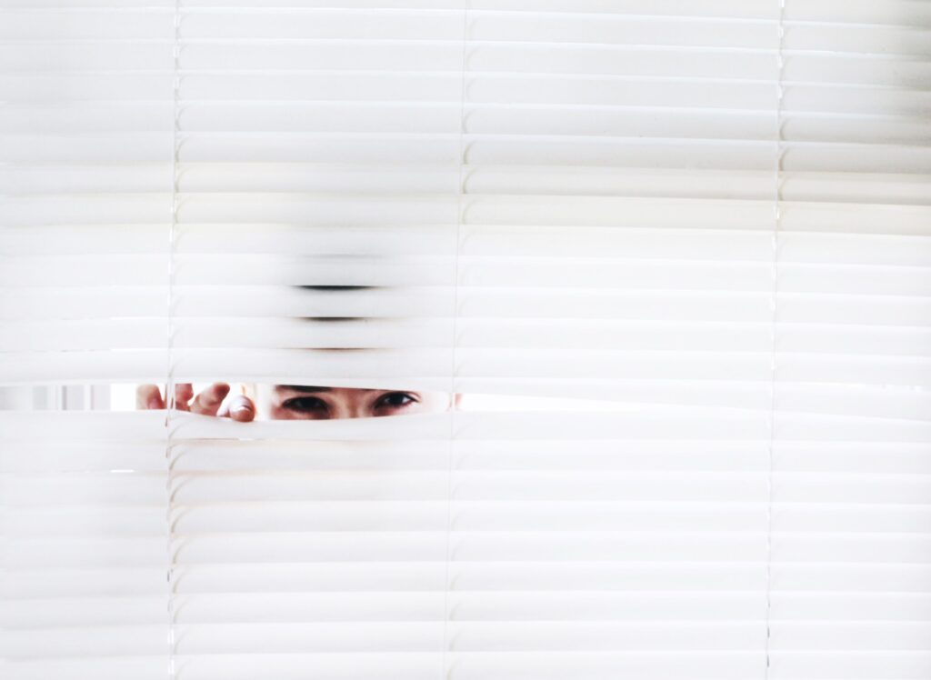 A person peeks through partially opened white window blinds, revealing only their eyes and hand while most of their face remains hidden behind the blinds. The background is bright and overexposed.