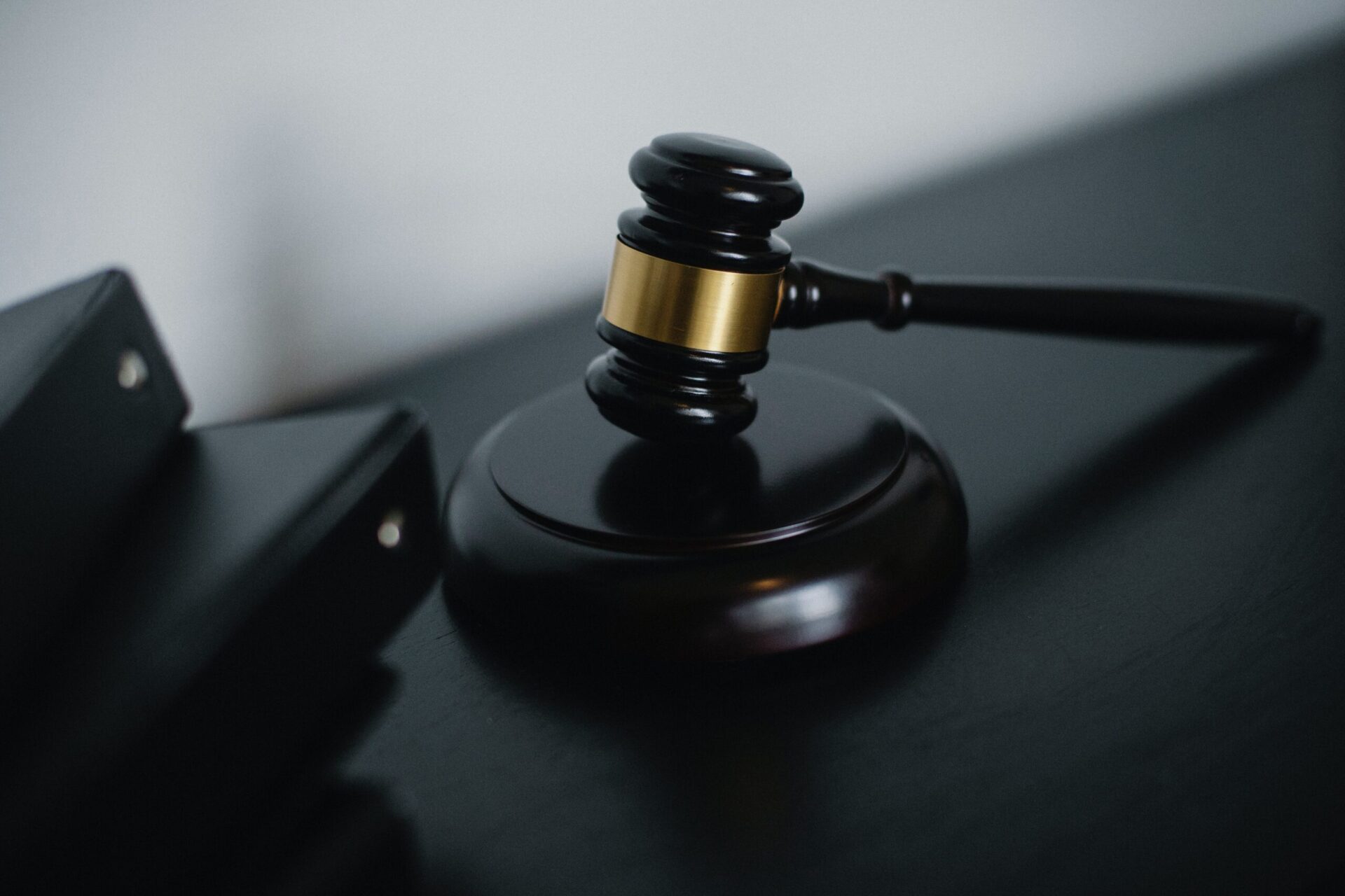 A close-up of a wooden judge’s gavel rests on a dark surface, with two black binders partially visible in the background. The  conveys a sense of law, justice, or legal proceedings.
