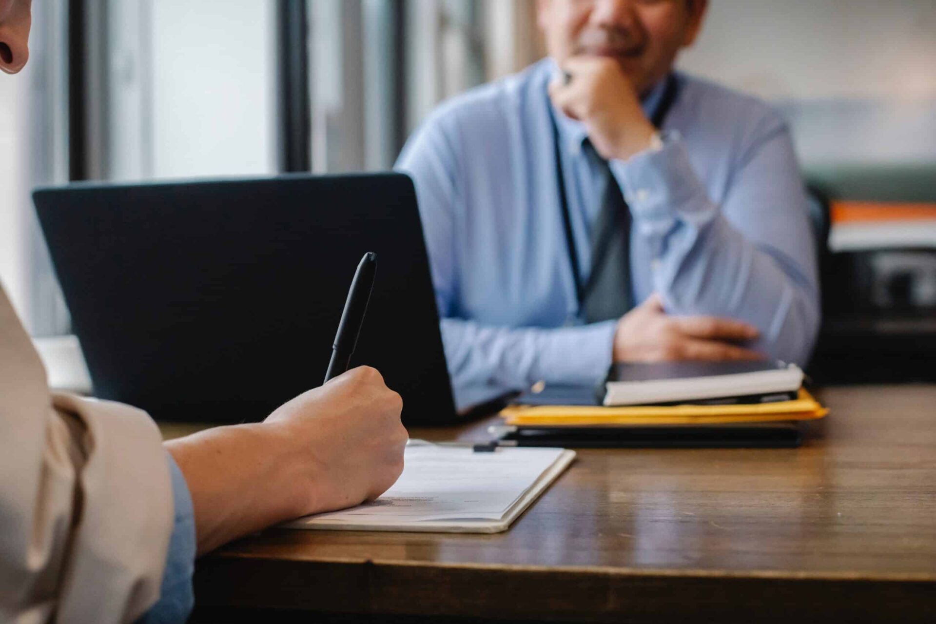 Two people sit at a wooden table during a meeting; one writes on a clipboard while the other, dressed in business attire, sits with a laptop and folders, listening attentively.