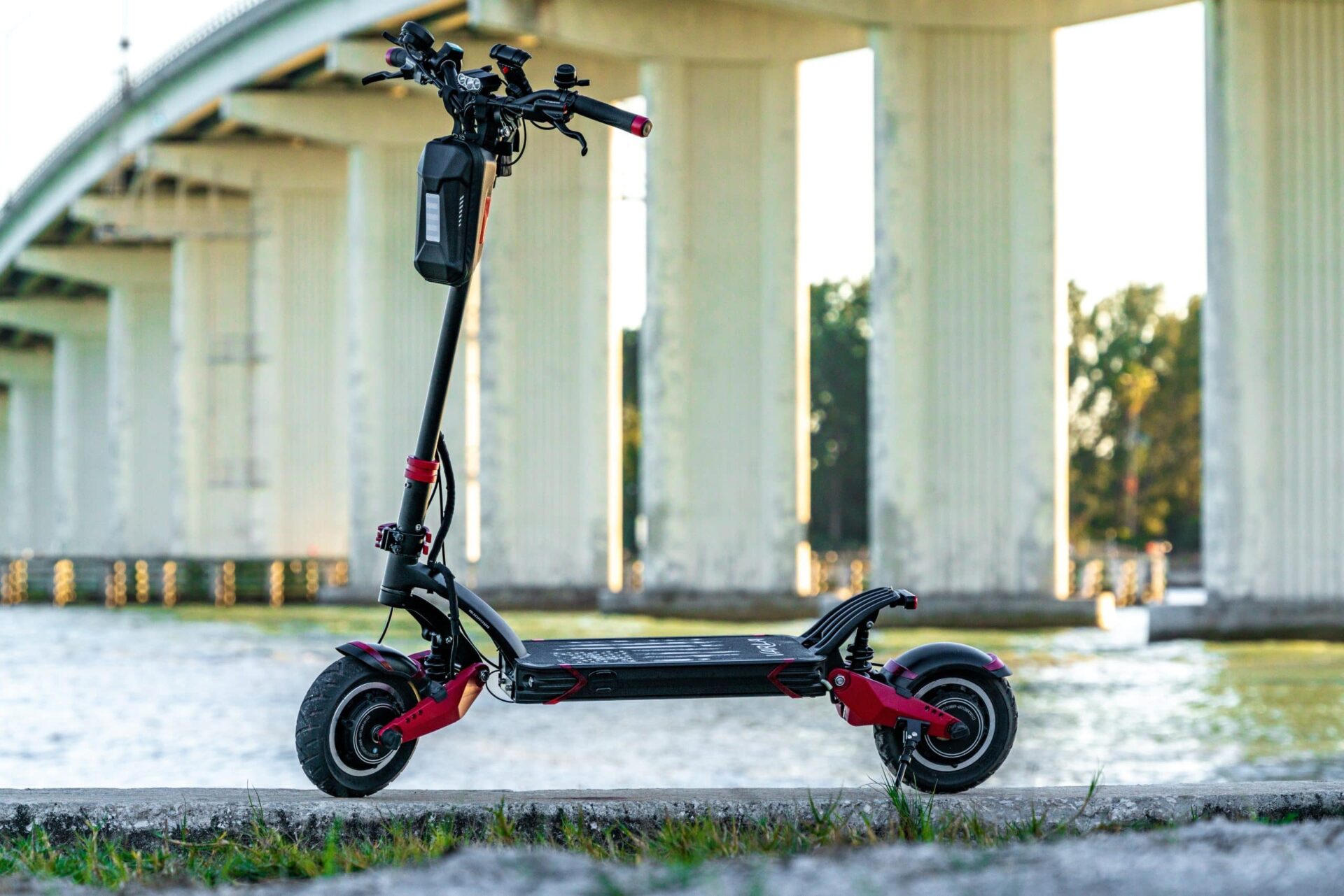 A black and red electric scooter parked on a concrete surface near water, with a large bridge and trees in the background.