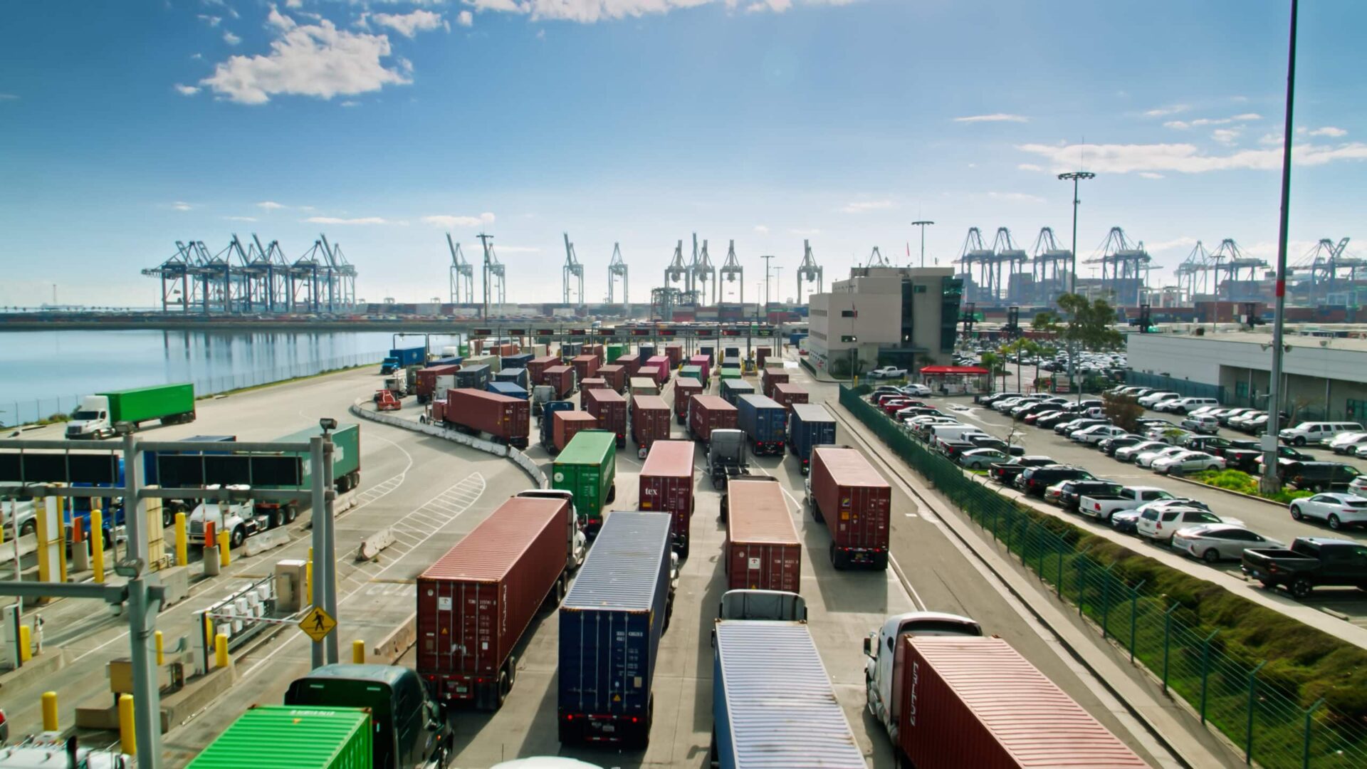 A busy port with rows of trucks carrying shipping containers, parked cars, and numerous cranes in the background under a bright blue sky.