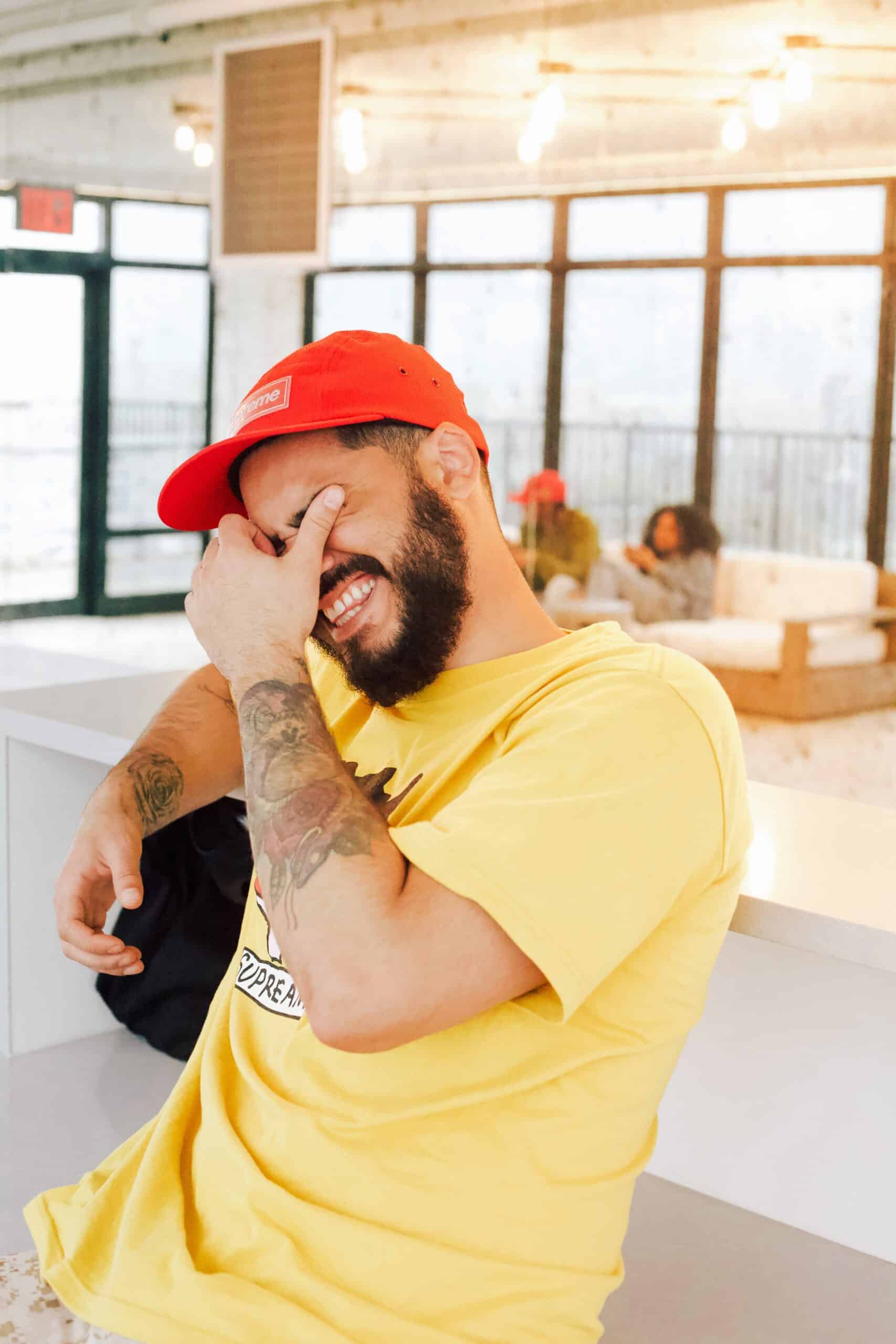 A man in a yellow t-shirt and red cap sits indoors, smiling and covering part of his face with his hand. He has tattoos on his arms. Blurred people and sofas are visible in the bright background.