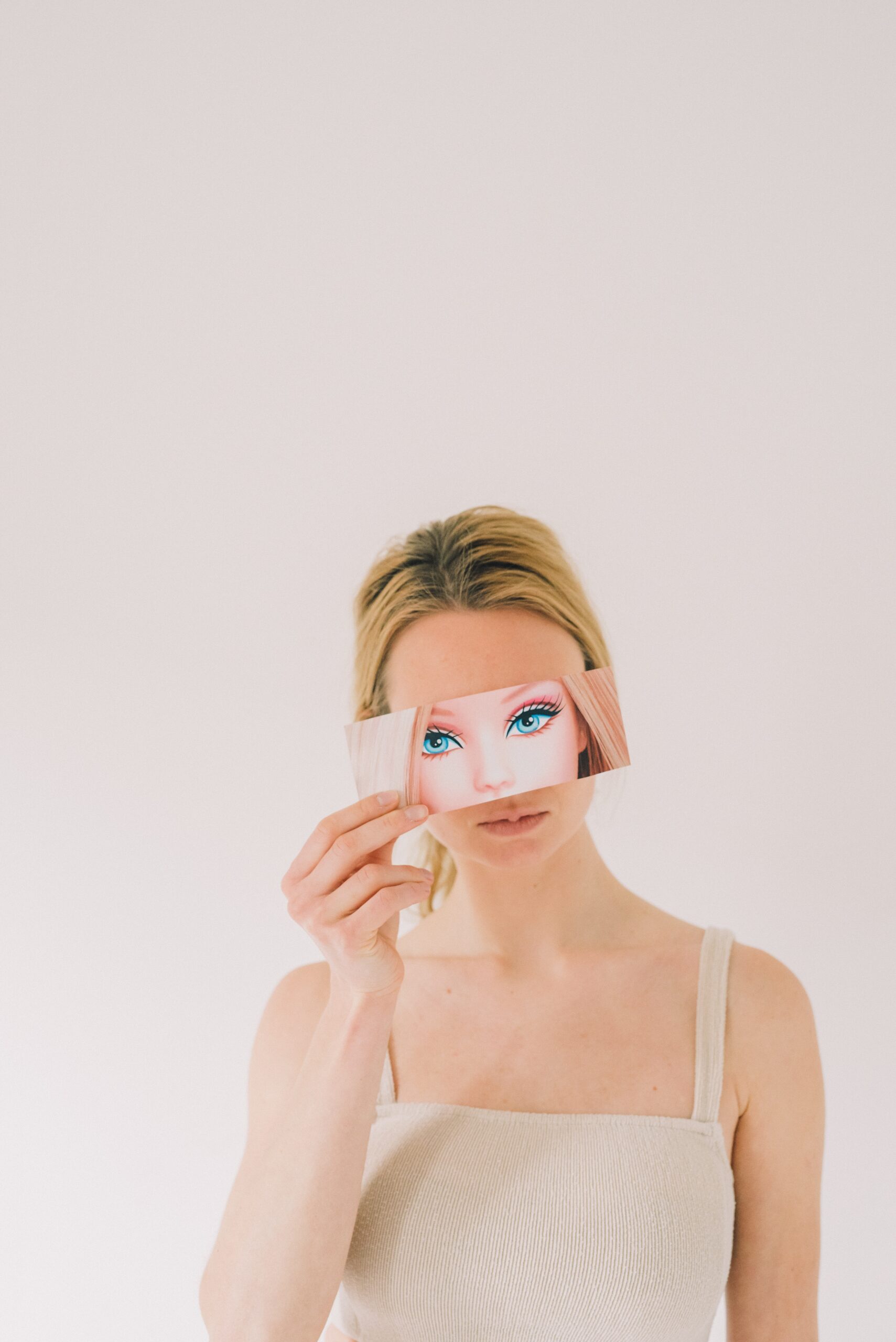 A woman wearing a beige tank top holds a cut-out  of a cartoon dolls eyes and hair in front of her face, aligning it with her own eyes, against a plain white background.