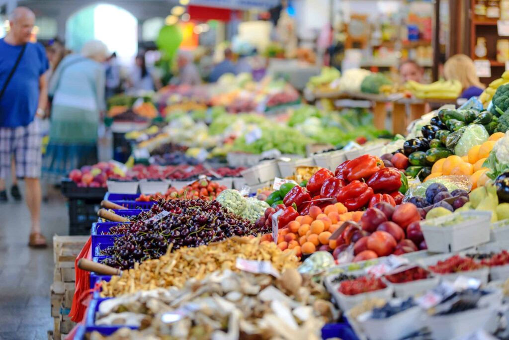 A bustling indoor market with colorful displays of fresh fruits, vegetables, and mushrooms. Shoppers walk among the stalls, browsing the abundant produce under bright lighting.