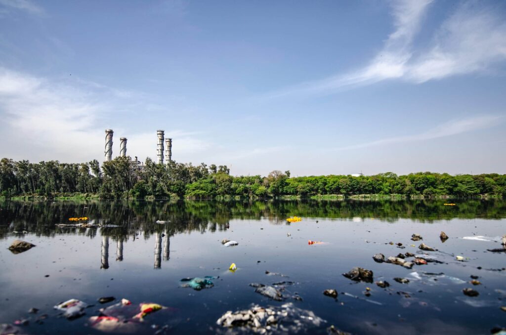 A polluted river with floating trash and debris, reflecting a factory and trees on the opposite bank under a blue sky with scattered clouds.