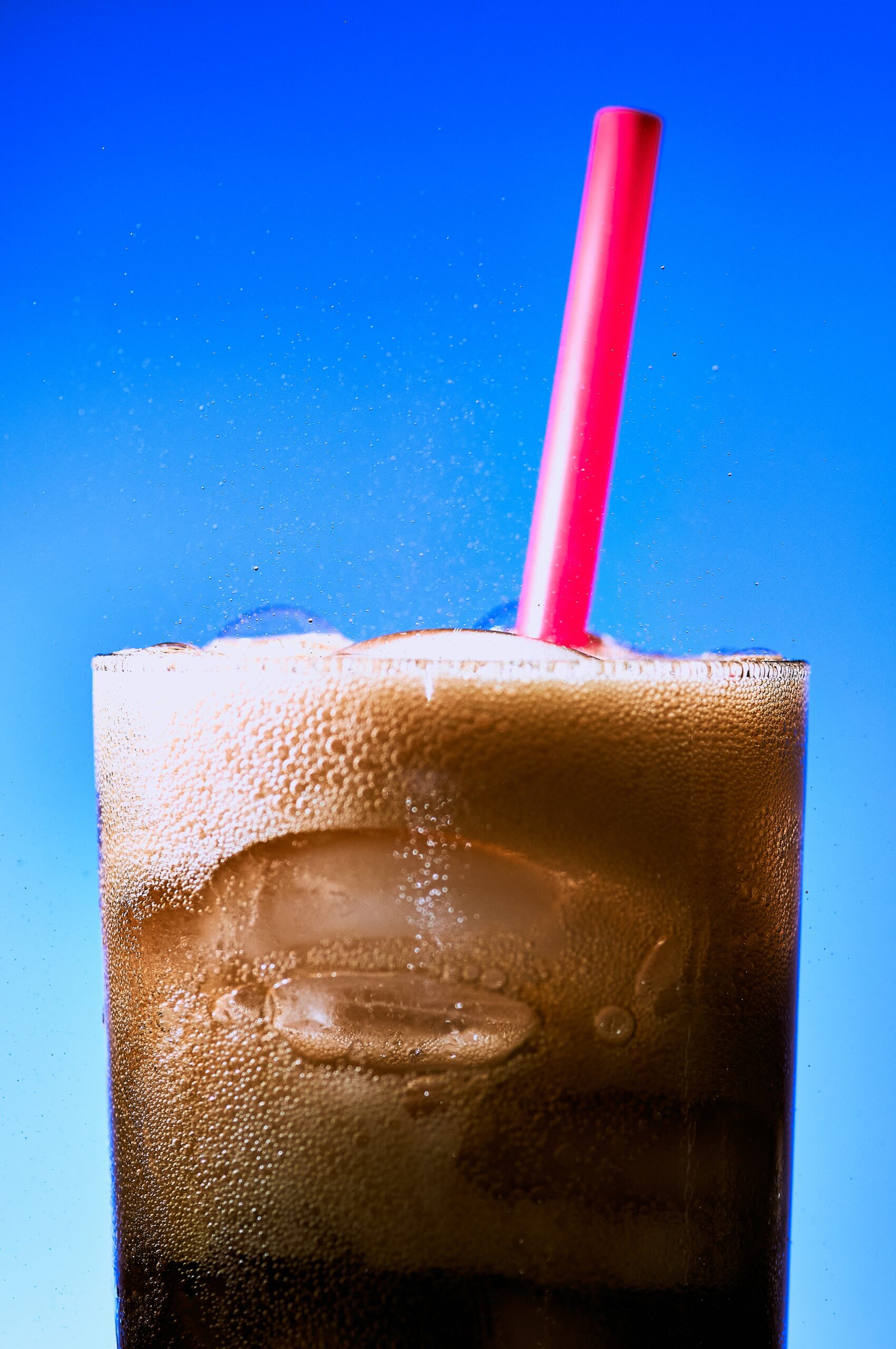 A close-up of a fizzy, iced dark soda in a glass with a red straw against a bright blue background. Bubbles and foam are visible on the surface of the drink.