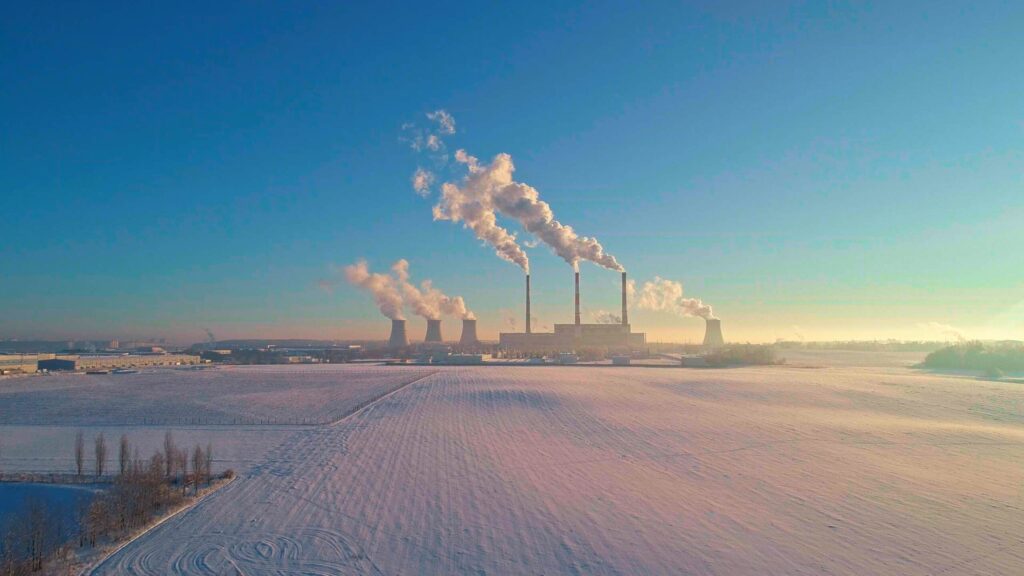 A power plant with multiple smokestacks emits white smoke over a vast, snow-covered landscape under a clear blue sky. The early morning sunlight casts a warm glow across the fields and industrial buildings.