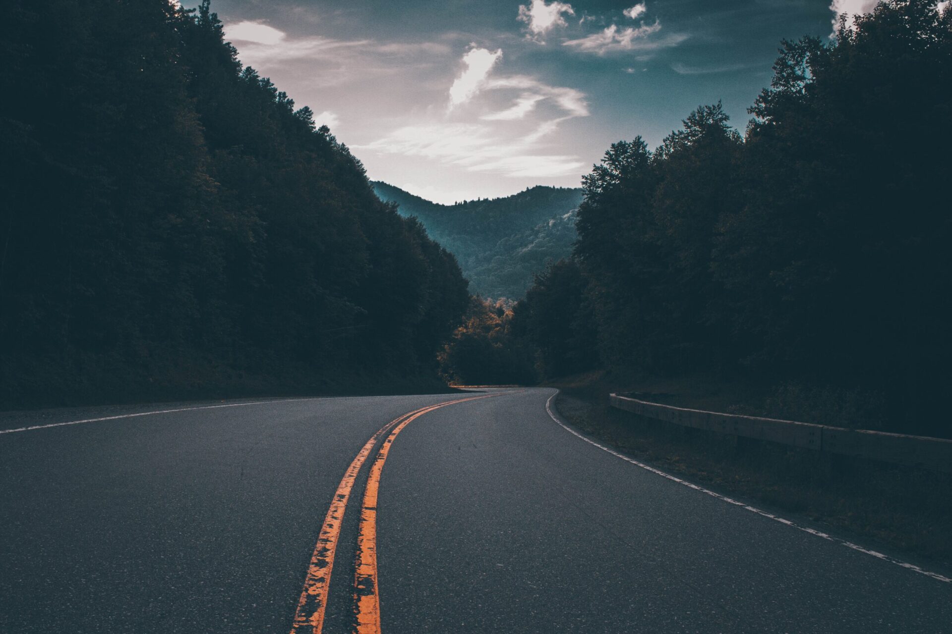 A winding road with a double yellow line curves through a forested area, bordered by trees, with mountains and a dramatic, cloudy sky in the background at dusk.