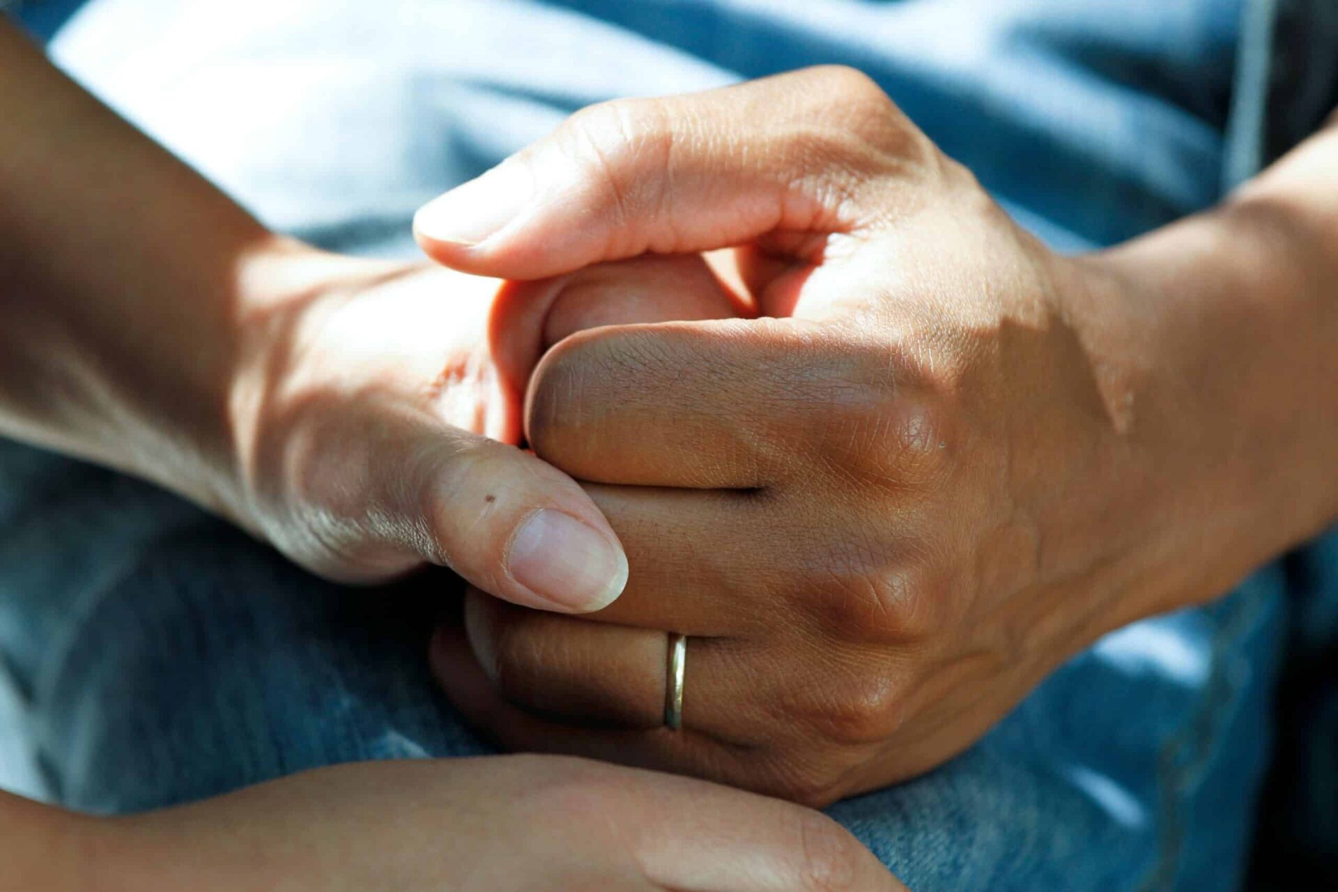 A close-up of two hands gently holding each other, one wearing a gold ring. The scene suggests comfort, support, or compassion, with soft lighting and a blue fabric background.