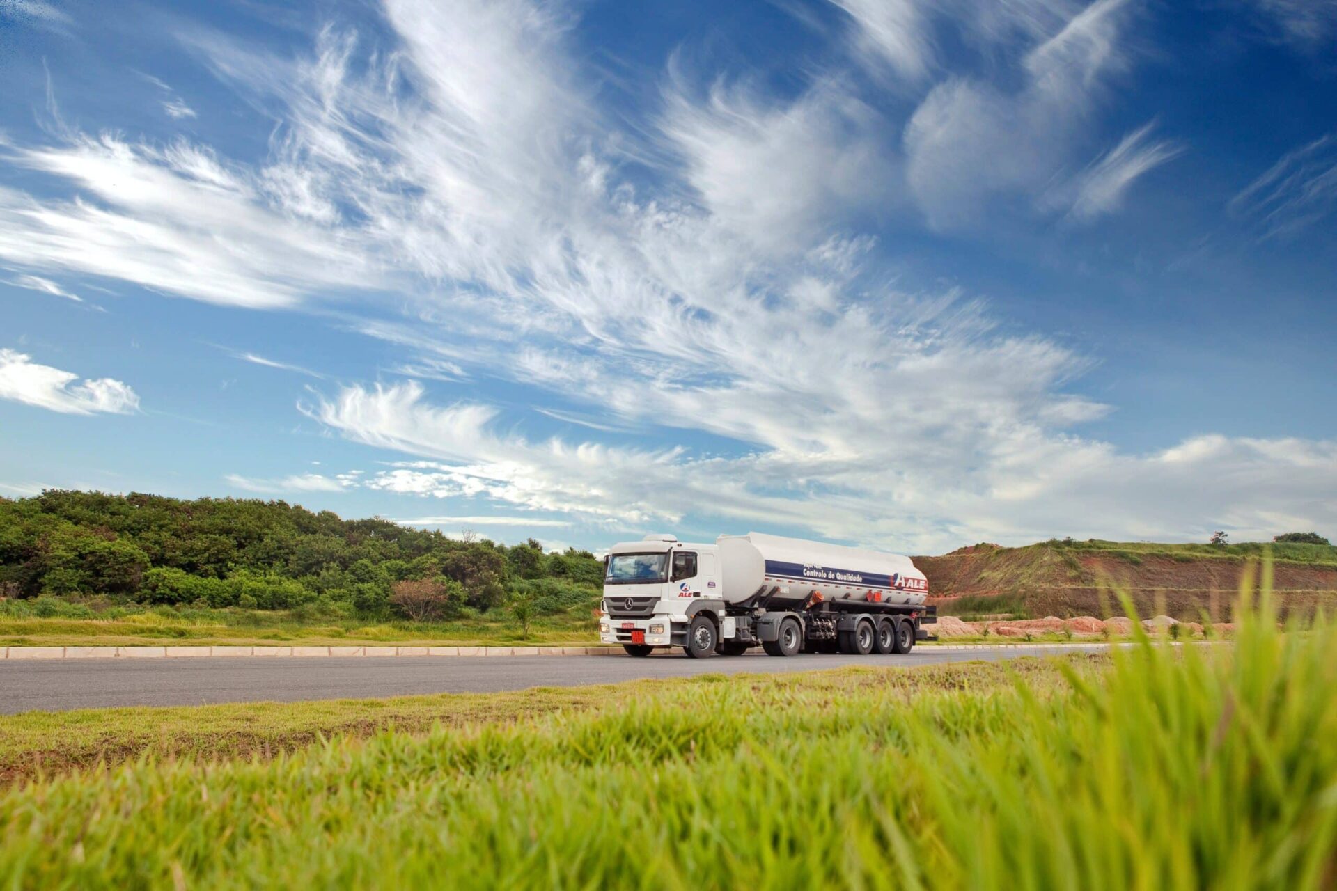 A large white fuel tanker truck drives on a paved road beside green grass and trees under a blue sky with wispy clouds.